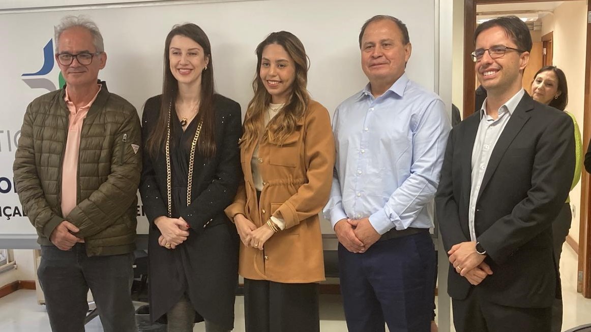 A imagem mostra cinco pessoas, três homens e duas mulheres, em pé lado a lado.

Da esquerda para a direita:

Um homem de cabelo grisalho e óculos, com um casaco bege sobre uma camisa polo rosada e calça escura.

Uma mulher de cabelo comprido e escuro, vestindo um blazer preto com detalhes dourados e uma saia preta.

Uma mulher de cabelo castanho claro e longo, com um casaco de tonalidade camel.

Um homem de camisa social azul clara e calça azul escura.

Um homem de óculos, blazer escuro, camisa clara e gravata. Ele tem as mãos cruzadas à frente.

Ao fundo, há uma parede branca com um logotipo institucional parcial visível no lado esquerdo.