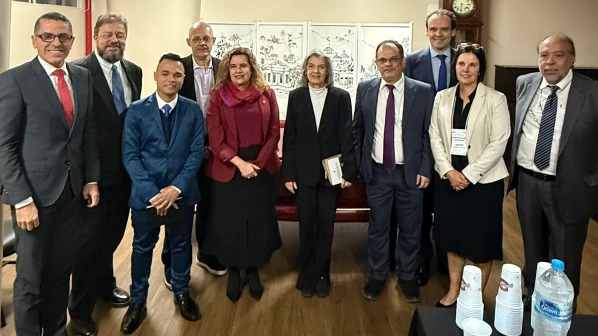 A imagem mostra um grupo de dez pessoas, a maioria homens e três mulheres, posando para uma foto em um ambiente interno.

Da esquerda para a direita:

Um homem de óculos, terno escuro, camisa branca e gravata vermelha.

Um homem com barba, terno escuro, camisa clara e gravata azul.

Um homem de terno azul, camisa azul e gravata escura, com as mãos cruzadas na frente.

Um homem careca, de terno cinza, camisa branca e gravata escura, com um pequeno bigode.

Uma mulher de casaco vinho sobre uma blusa clara, saia preta e sapatos escuros.

Uma mulher com cabelos grisalhos e longos, vestindo um casaco escuro sobre uma blusa branca. Ela segura algo nas mãos.

Um homem de terno azul escuro, camisa branca e gravata roxa.

Um homem sorrindo, de terno azul escuro, camisa branca e gravata azul.

Uma mulher de blazer branco sobre uma blusa preta, com um crachá de identificação no lado direito.

Um homem com bigode, terno escuro, camisa branca e gravata listrada.