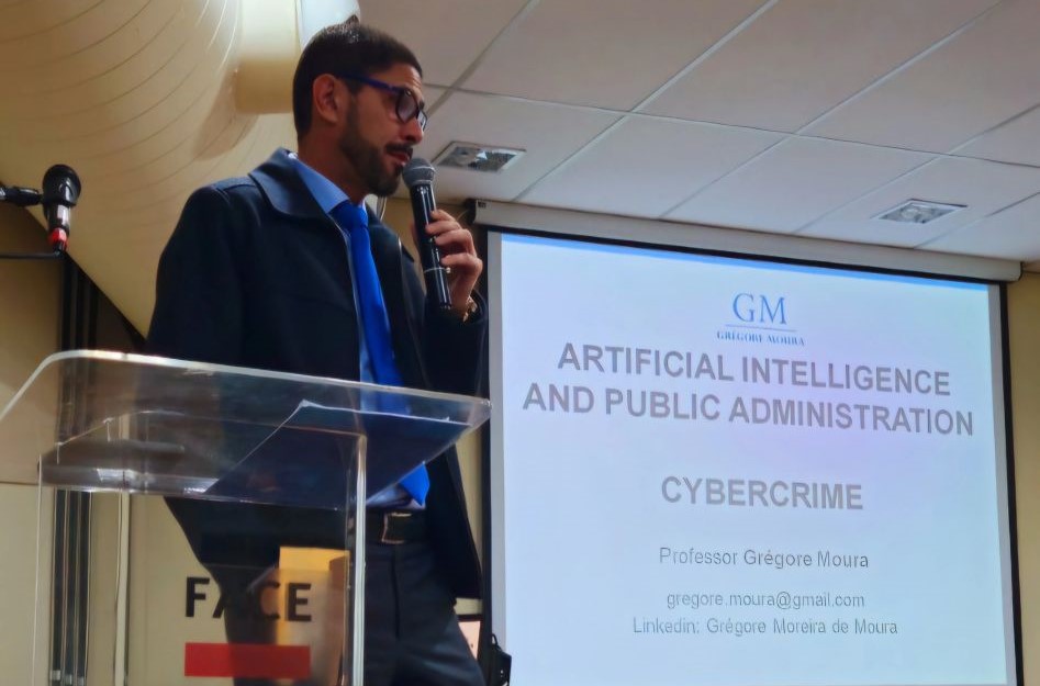 A imagem mostra um homem com óculos e barba, vestindo uma camisa de botão e gravata, falando em um microfone. Ele está em pé atrás de um púlpito com um logo. Atrás dele, há uma tela de projeção com o texto "GM - ARTIFICIAL INTELLIGENCE AND PUBLIC ADMINISTRATION", "CYBERCRIME" e o nome do palestrante, "Professor Grégore Moura", junto com seu e-mail e perfil do LinkedIn.