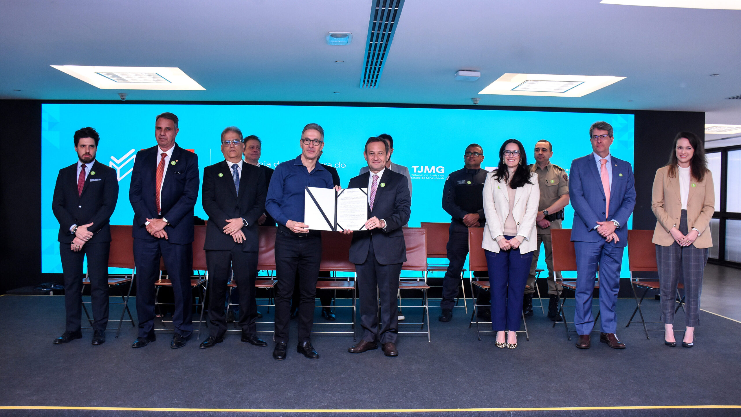 A imagem mostra um grupo de oito pessoas lado a lado em pé em frente a um grande telão azul. No centro da imagem, dois homens estão de frente um para o outro, segurando um documento assinado. Os outros estão em pé ao redor deles. Todos estão vestidos com roupas formais. Há um pequeno grupo de cadeiras dispostas em fileira atrás do grupo de pessoas.