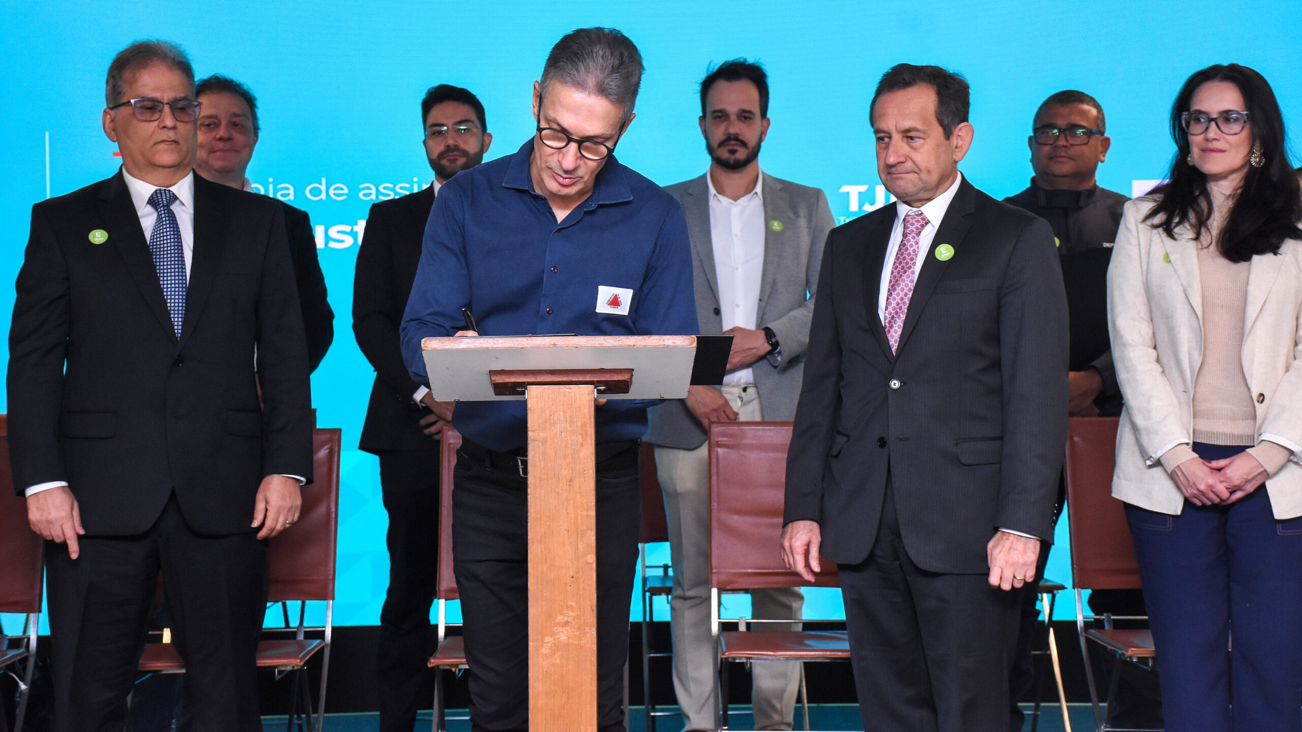 A imagem mostra um grupo de oito pessoas, sete homens e uma mulher, em um evento. Um homem no centro, vestindo uma camisa azul-marinho, está em pé em um púlpito, assinando um documento. Ele usa óculos e tem cabelos grisalhos. Ao lado e atrás dele, há outros homens e a mulher em pé, observando. Todos estão vestidos com roupas formais, e o fundo é uma parede azul com letras brancas parcialmente visíveis.
