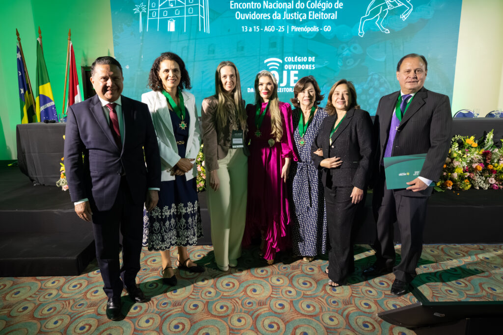 A imagem mostra um grupo de sete pessoas, dois homens e cinco mulheres, em um evento formal. No centro, uma mulher de vestido rosa-escuro. Ao seu redor, as outras pessoas estão posando para a foto. Todos estão com roupas formais e a maioria usa uma faixa verde no pescoço. O fundo tem um telão com o logo "COJE" e tem algumas bandeiras hasteadas.