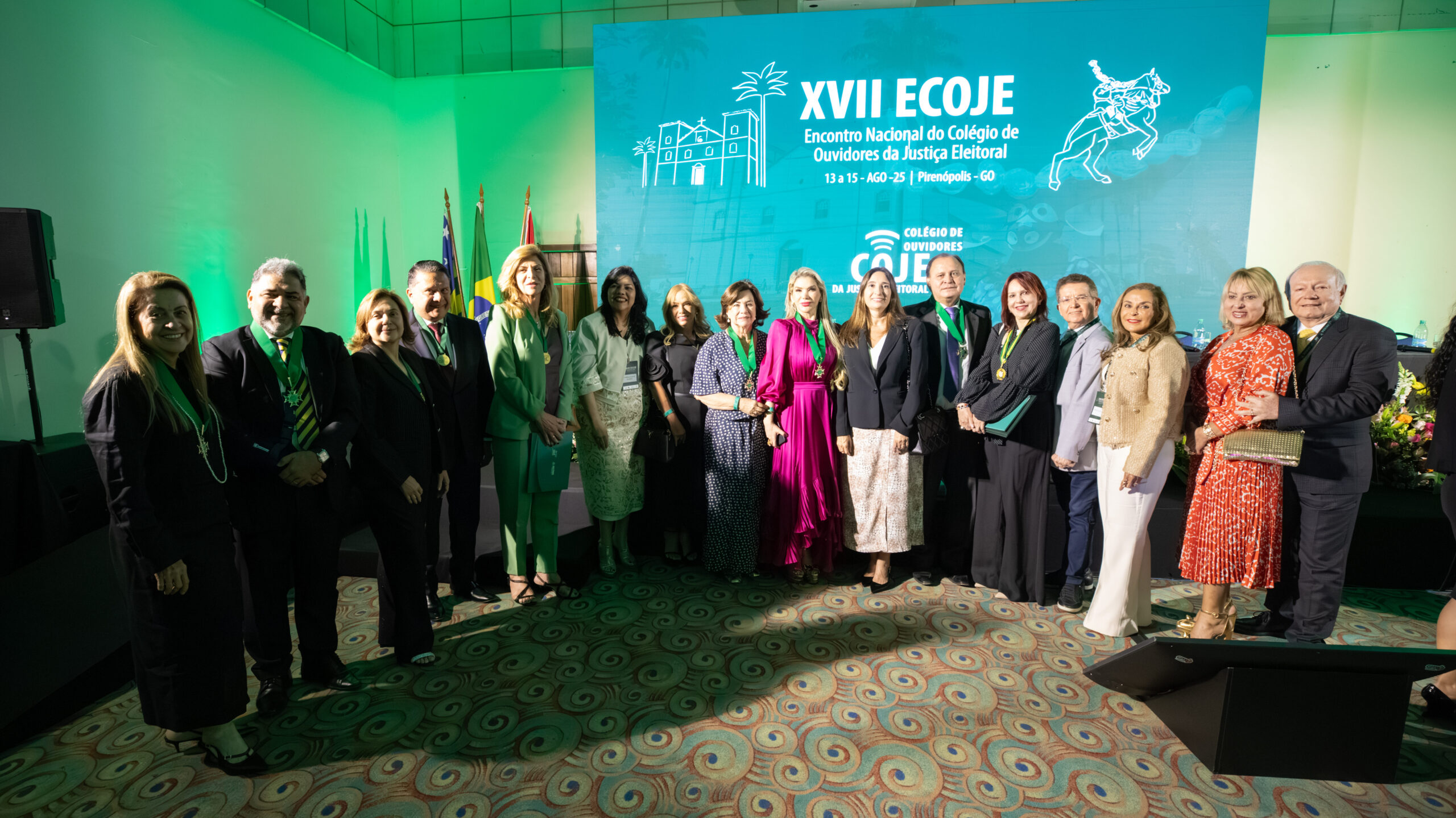A imagem mostra um grande grupo de pessoas, homens e mulheres, em um ambiente de conferência. Eles estão todos de pé, posando para uma foto em grupo, em frente a um grande painel azul-claro com a inscrição "XVII ECOJE Encontro Nacional do Colégio de Ouvidores da Justiça Eleitoral". O ambiente está iluminado com luzes verdes.