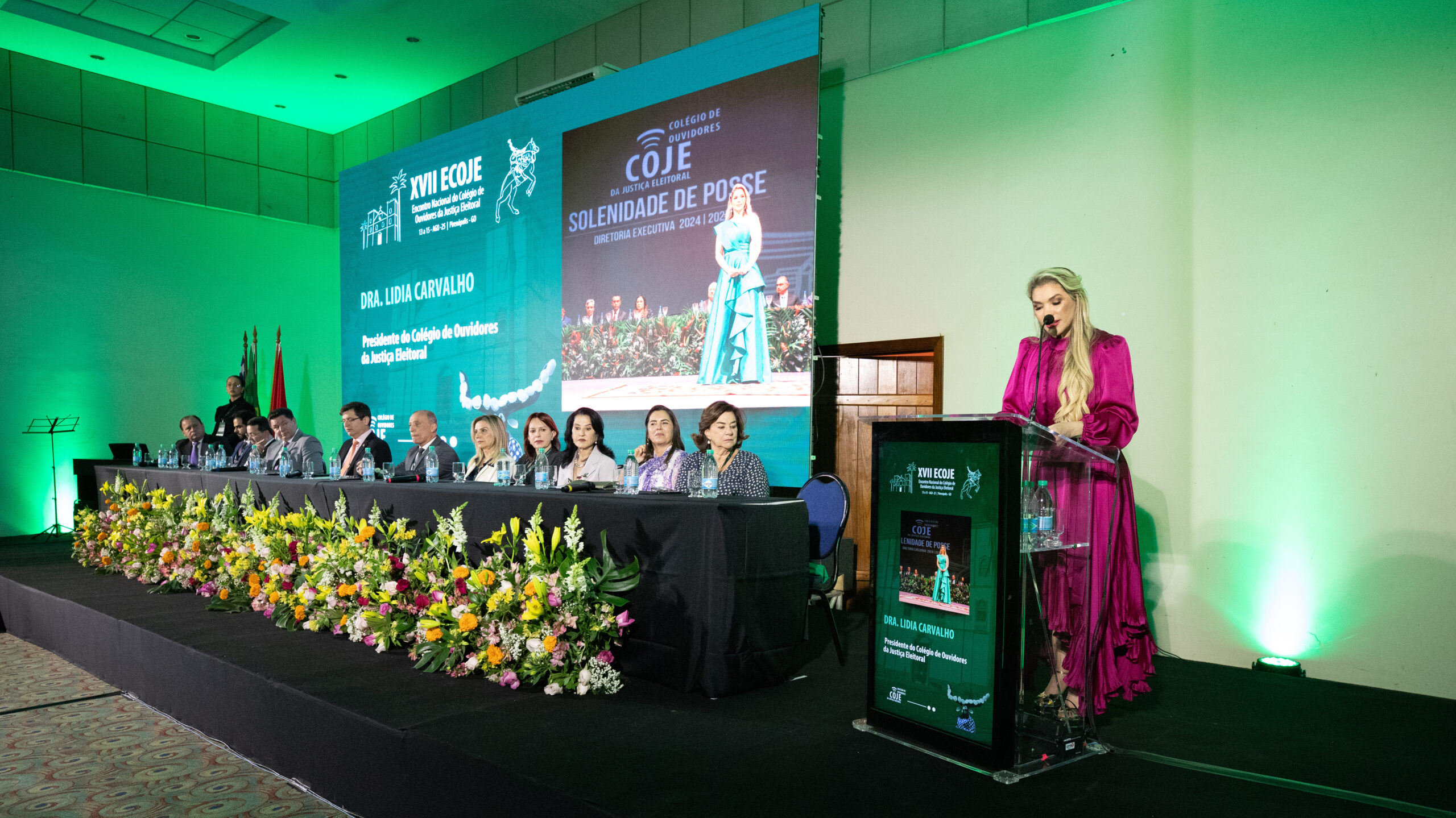 A imagem mostra uma mulher de cabelos loiros e vestido rosa-escuro, em pé, falando em um púlpito. Ela está em um palco, onde há uma mesa longa com nove pessoas sentadas atrás dela. Na frente da mesa, há um arranjo de flores. Ao fundo, um grande telão exibe o logo do "COJE - SOLENIDADE DE POSSE". Há também outro telão menor no púlpito com o texto "DRA. LIDIA CARVALHO" e "Presidente do Colégio de Ouvidores da Justiça Eleitoral". O ambiente está iluminado com luzes verdes.