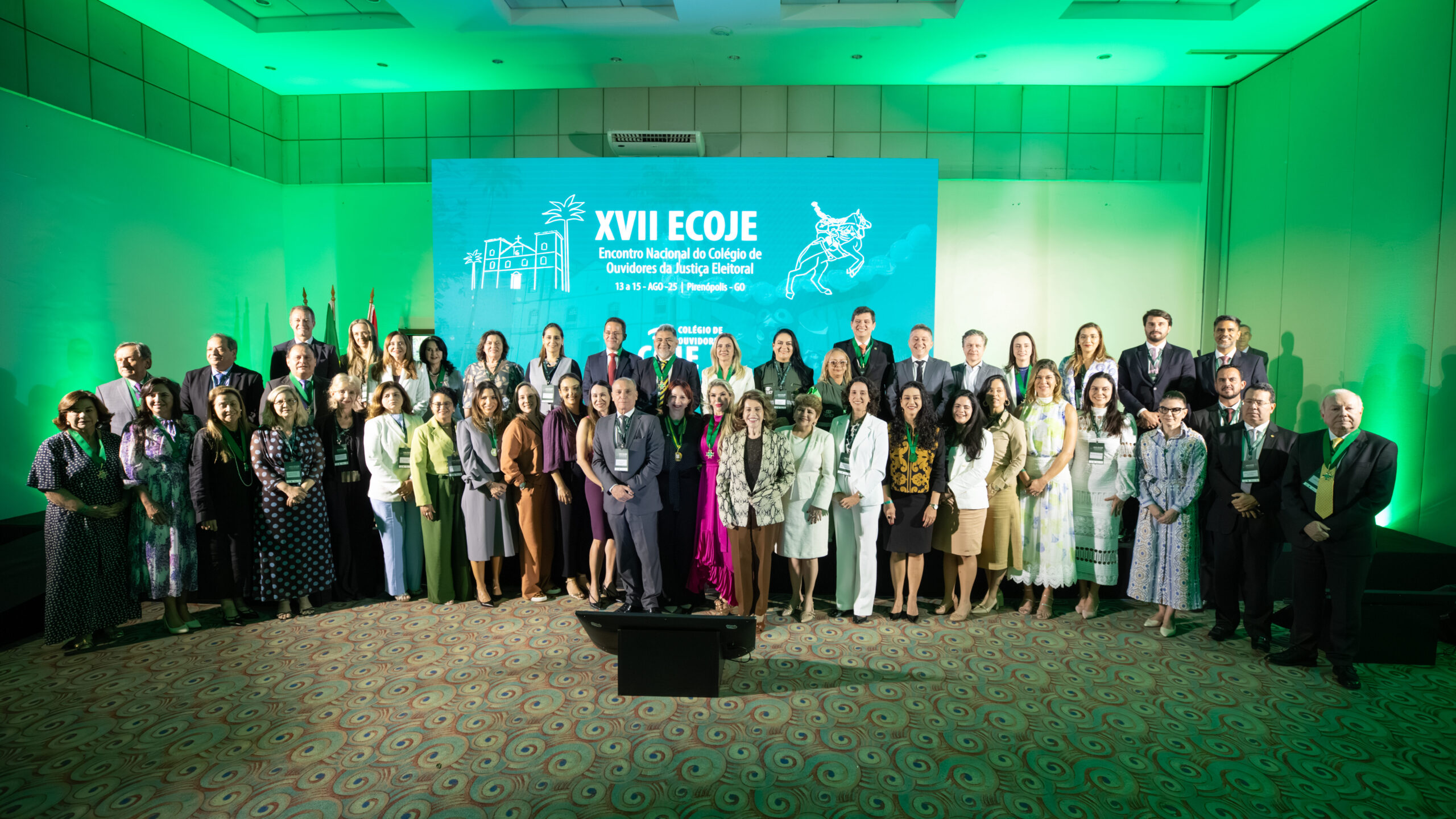 A imagem mostra um grande grupo de pessoas, homens e mulheres, em um ambiente de conferência. Eles estão todos de pé, posando para uma foto em grupo, em frente a um grande painel azul-claro com a inscrição "XVII ECOJE Encontro Nacional do Colégio de Ouvidores da Justiça Eleitoral". O ambiente está iluminado com luzes verdes.