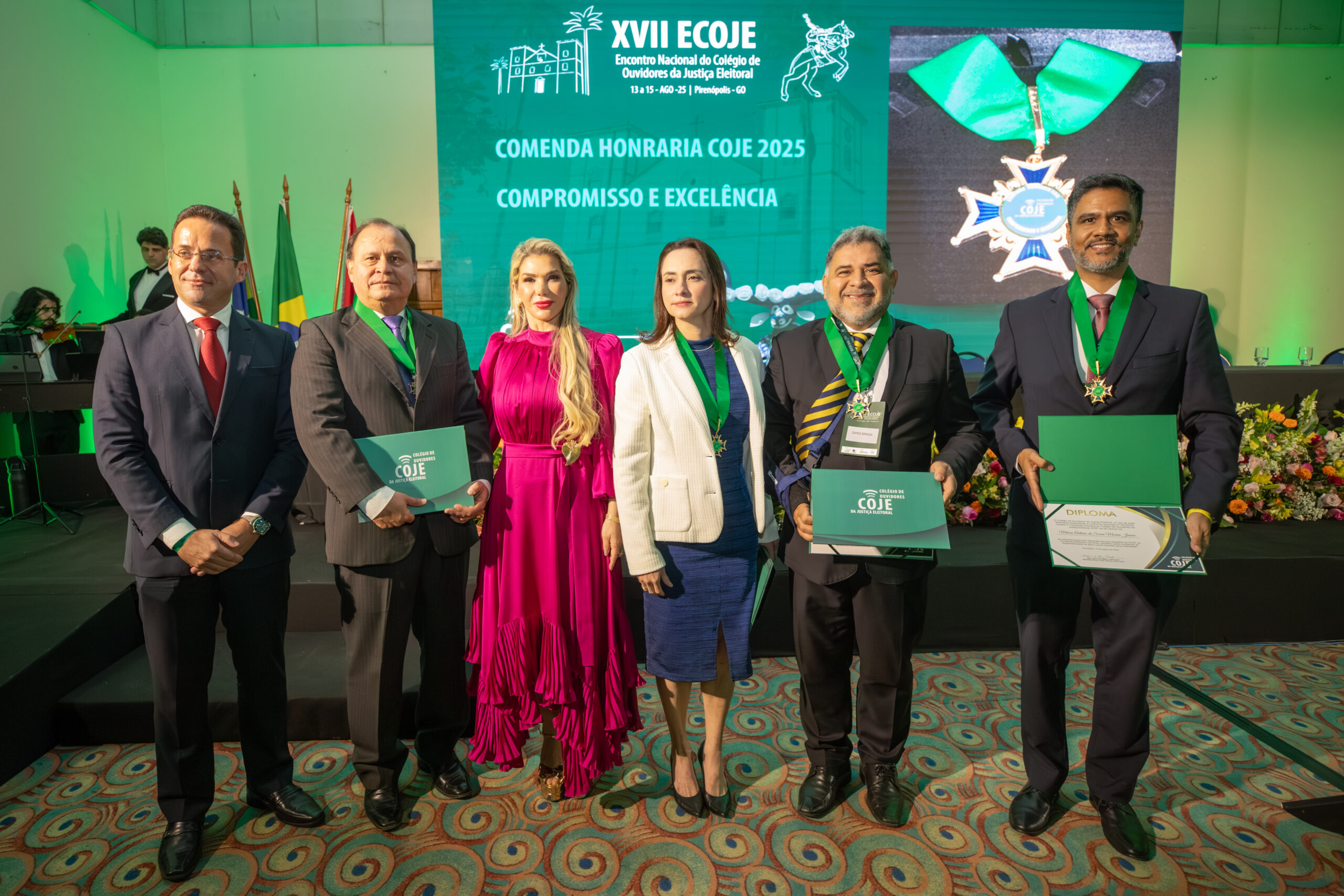 A imagem mostra seis pessoas, quatro homens e duas mulheres, todos vestidos em trajes formais, em um evento de premiação. Eles estão em pé, posando para uma foto em frente a um grande painel verde.
No centro do painel, o texto "COMENDA HONRARIA COJE 2025 COMPROMISSO E EXCELÊNCIA". No canto superior esquerdo, o logo do evento com a inscrição "XVII ECOJE".