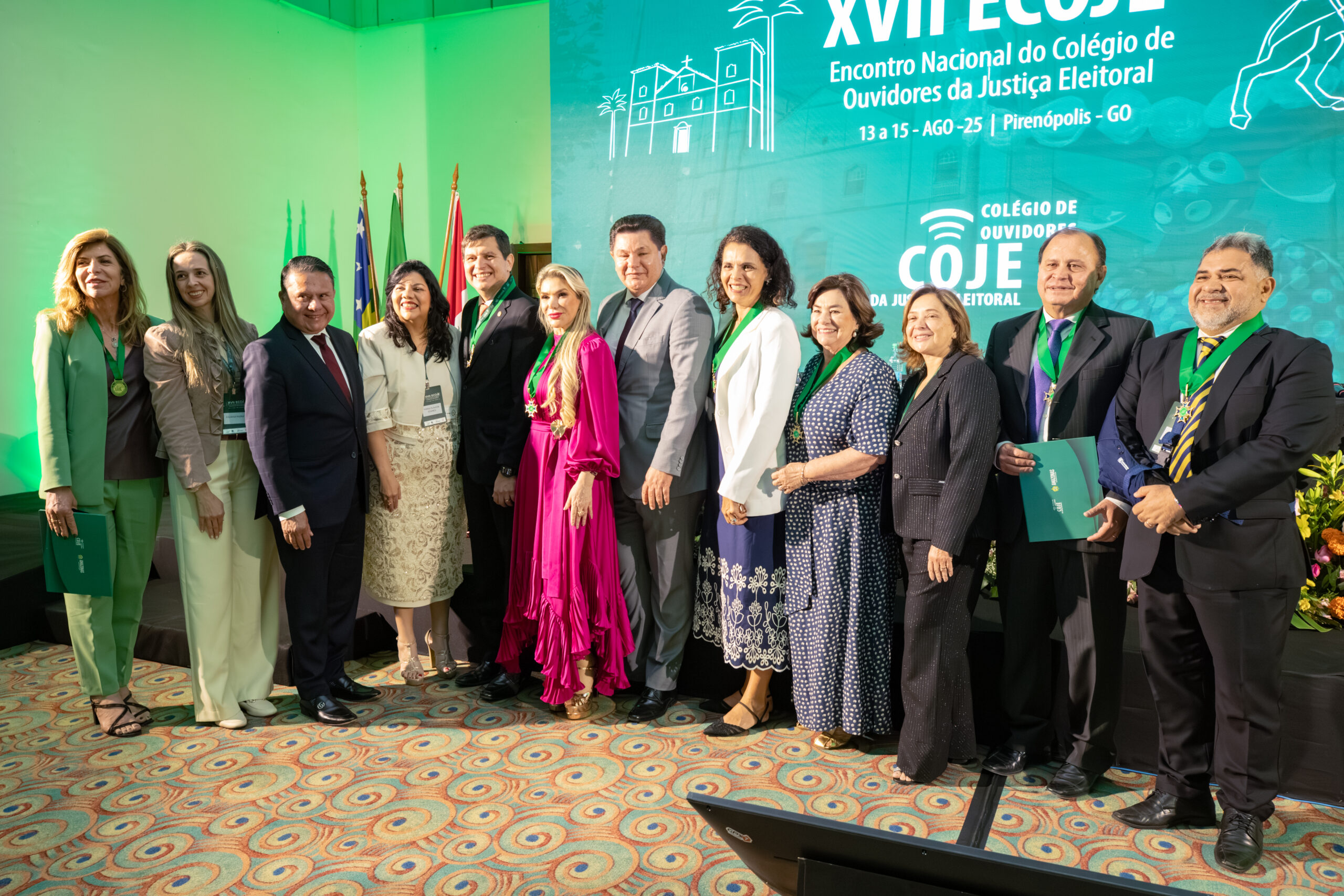 A imagem mostra um grande grupo de pessoas em um evento formal. Eles estão em pé, posando para uma foto em grupo, em frente a um grande painel azul-claro com a inscrição "XVII ECOJE Encontro Nacional do Colégio de Ouvidores da Justiça Eleitoral". A maioria está com uma faixa verde no pescoço. O ambiente está iluminado com luzes verdes.