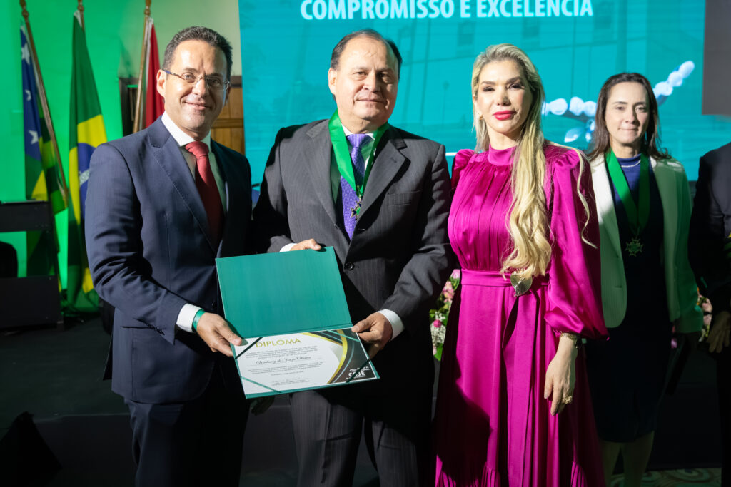 A imagem mostra um grupo de quatro pessoas, dois homens e duas mulheres, em um evento formal. No centro, um homem com terno escuro e uma faixa verde no pescoço está segurando um diploma. Ao lado dele, um homem com terno azul-marinho segura o diploma. Uma mulher de vestido rosa, com cabelos loiros e compridos, e outra mulher com cabelos escuros, também com uma faixa verde, estão ao lado deles. O grupo está posando para uma foto em frente a um painel verde-claro com a inscrição "COMPROMISSO E EXCELÊNCIA".