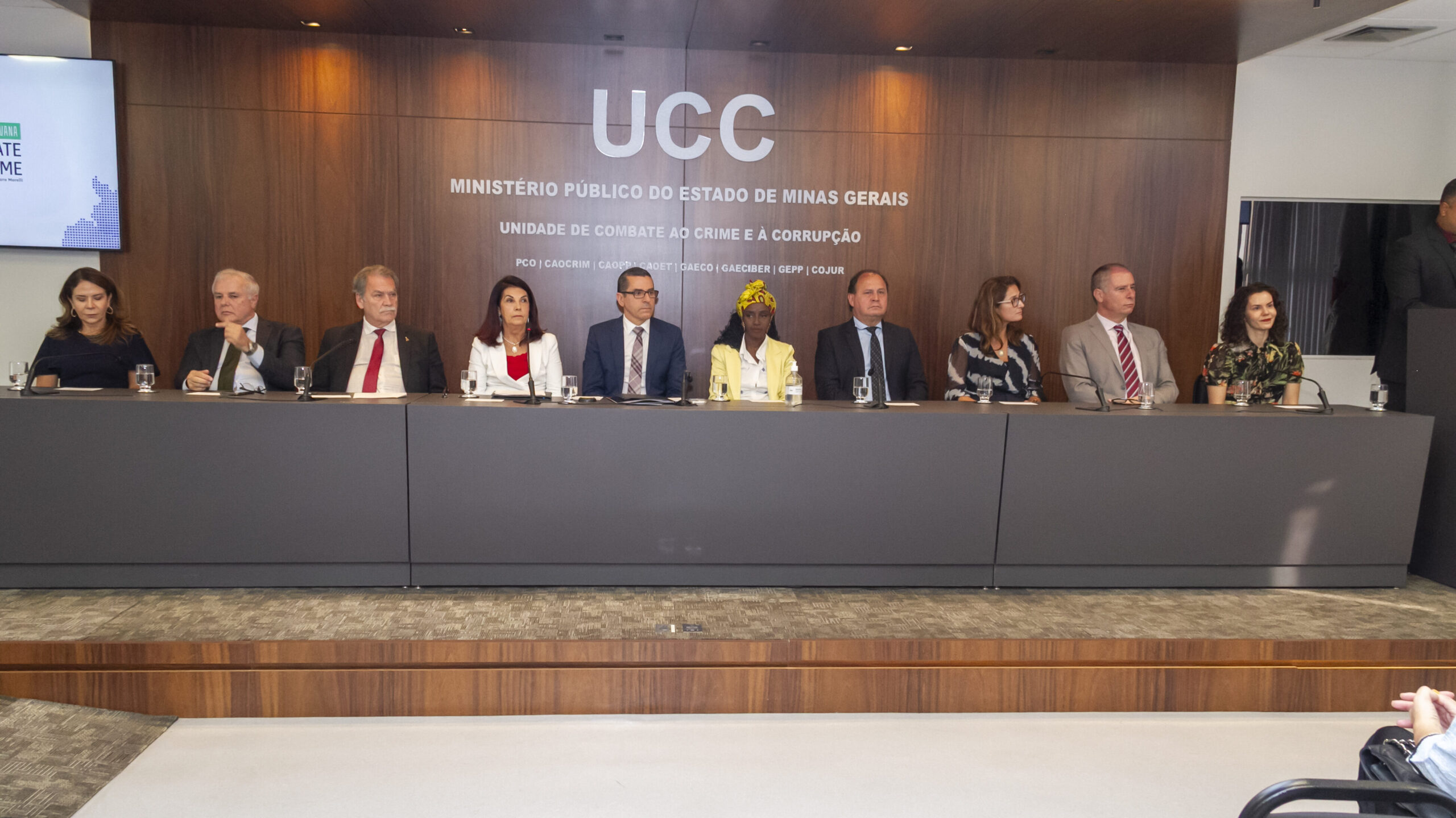 A imagem mostra dez pessoas, incluindo homens e mulheres, sentadas em uma mesa de painel. Eles estão em frente a uma parede de madeira escura, que exibe os logotipos "UCC" e o texto "MINISTÉRIO PÚBLICO DO ESTADO DE MINAS GERAIS", junto com o subtítulo "UNIDADE DE COMBATE AO CRIME E À CORRUPÇÃO".
Todos estão vestidos de forma formal e parecem ser autoridades ou palestrantes. Há copos de água sobre a mesa à frente deles. À esquerda, um telão pode ser visto.