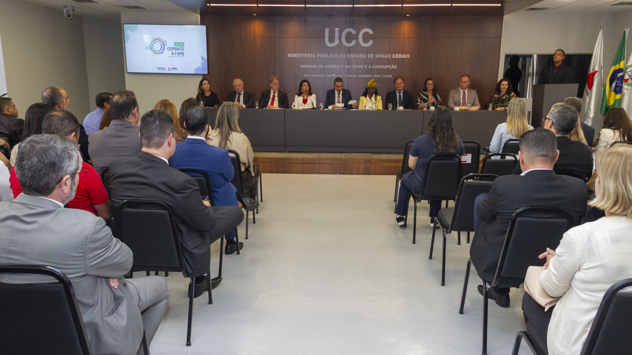 A imagem mostra uma reunião ou evento em um auditório. Em primeiro plano, é possível ver a parte de trás de várias pessoas sentadas em fileiras de cadeiras pretas, de costas para a câmera. Em segundo plano, em uma mesa de madeira escura, há dez pessoas sentadas.
Acima deles, em uma parede com a mesma madeira, há um logotipo e o texto "UCC MINISTÉRIO PÚBLICO DO ESTADO DE MINAS GERAIS". À direita, um homem está discursando em um púlpito.