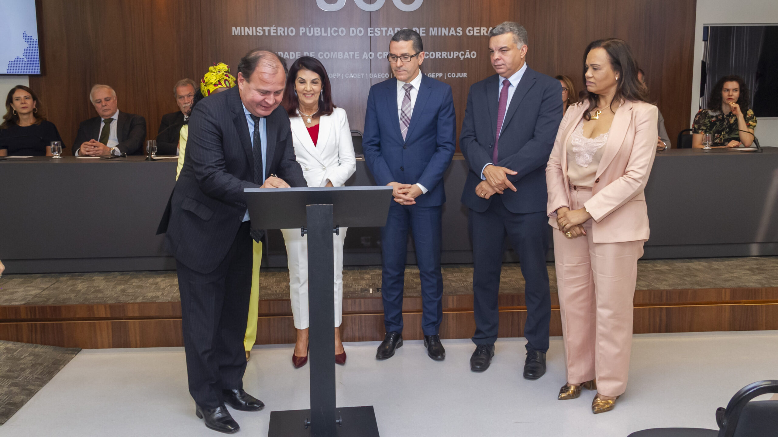 A imagem mostra uma cerimônia de assinatura em um evento interno. O foco está em cinco pessoas. Um homem de terno escuro e gravata está assinando um documento em um pódio. Atrás e ao lado dele, outras quatro pessoas estão em pé.
Da esquerda para a direita, atrás do homem que assina, estão: uma mulher de blazer branco e blusa vermelha; um homem com óculos e terno azul; um homem com terno escuro; e uma mulher vestindo um conjunto de blazer e calça social em tom de rosa-claro. Outras pessoas podem ser vistas ao fundo, sentadas em uma mesa, em um ambiente formal. A placa no pódio indica "MINISTÉRIO PÚBLICO DO ESTADO DE MINAS GERAIS".