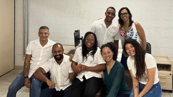 A imagem mostra um grupo de sete pessoas, quatro mulheres e três homens, em um ambiente interno. Eles estão todos de pé ou sentados, sorrindo para a câmera. Quase todos estão vestindo camisetas brancas. A maioria das pessoas está sentada, enquanto duas pessoas estão de pé atrás.