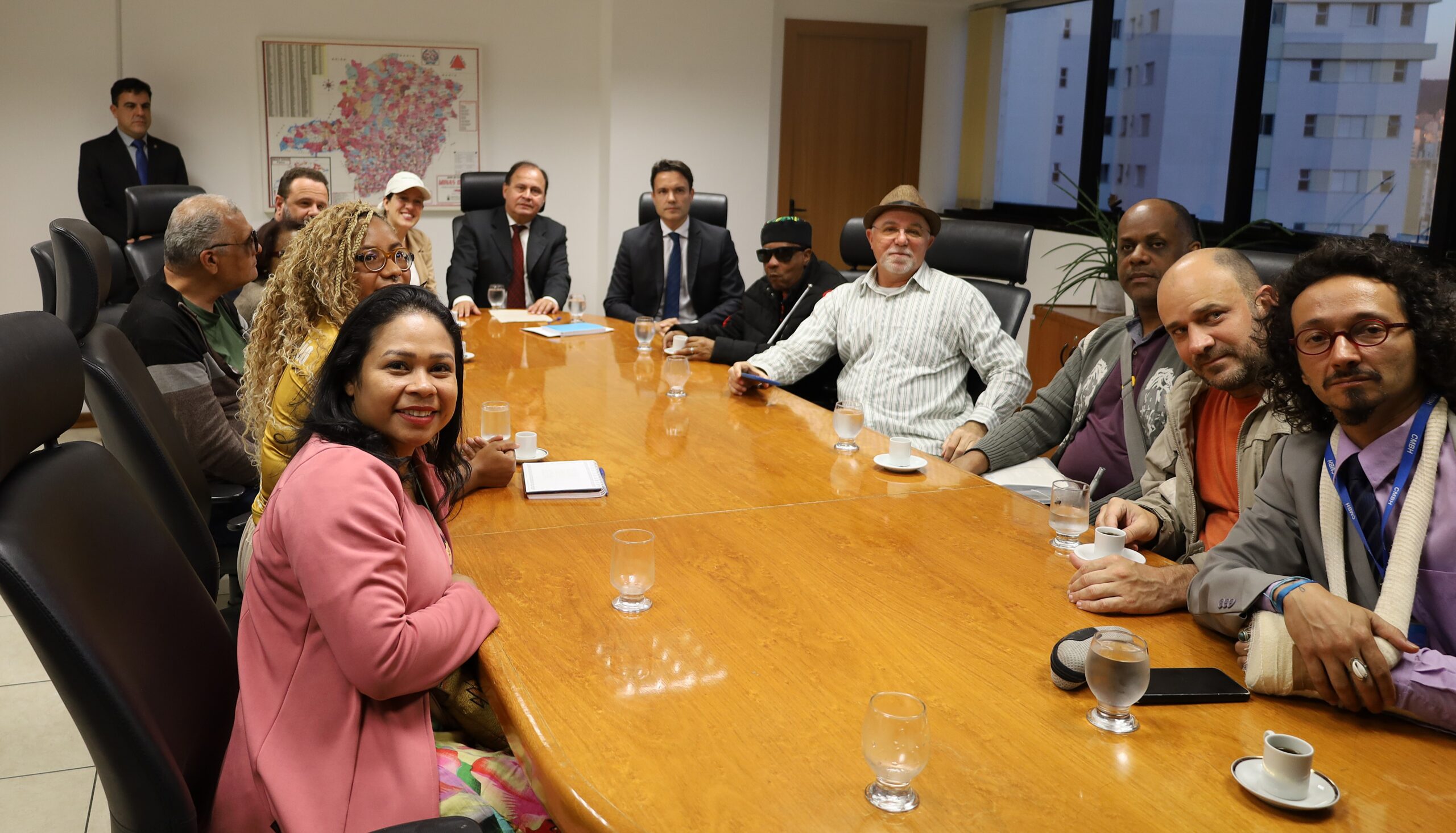 A imagem mostra um grupo de pessoas sentadas ao redor de uma grande mesa de madeira. Eles estão em uma sala de reunião. Ao fundo, uma mapa de Minas Gerais e algumas janelas são visíveis.