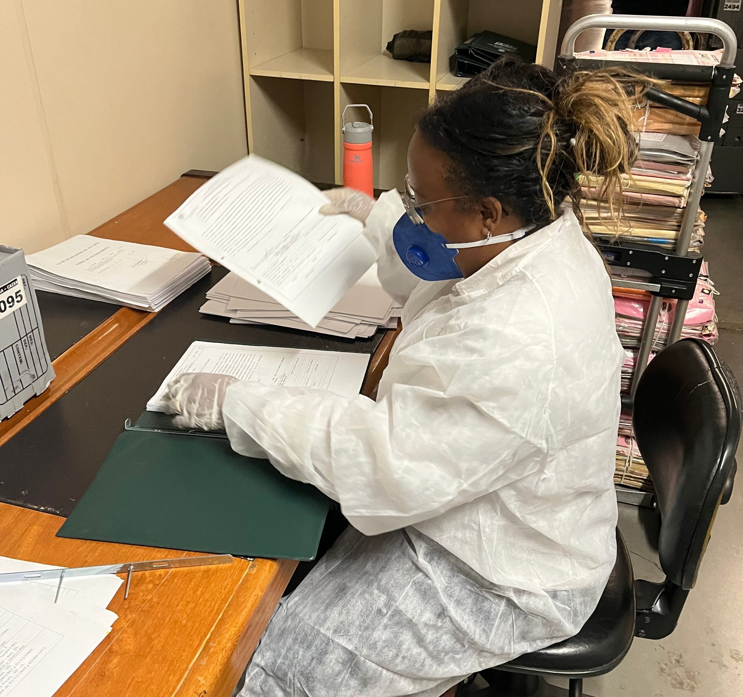 A imagem mostra uma mulher sentada em uma mesa. Ela está usando uma máscara de proteção azul, luvas brancas e um jaleco branco sobre a roupa. A mulher está manuseando vários documentos de papel, que estão sobre a mesa. Ao fundo, há prateleiras e um carrinho com pilhas de papéis.