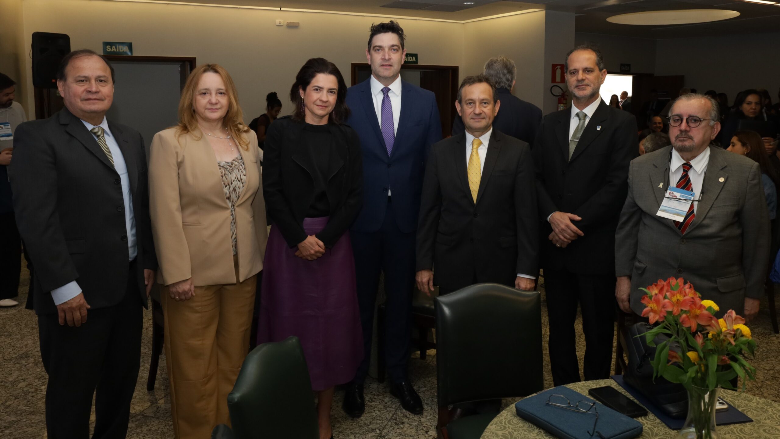 A imagem mostra sete pessoas, cinco homens e duas mulheres, em pé, posando para uma foto em um evento. Eles estão vestidos com trajes formais de trabalho. Em primeiro plano, no canto inferior direito, há uma mesa com um vaso de flores.