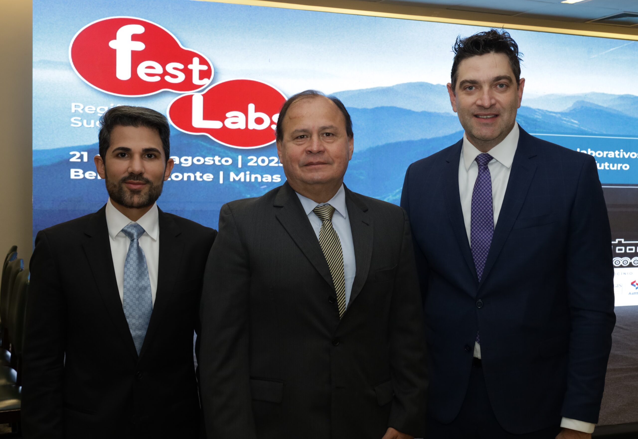 A imagem mostra três homens, todos de terno e gravata, posando para uma foto em um evento.
O homem da esquerda tem barba e cabelo escuros, com um terno preto e gravata azul. O homem do centro tem cabelo escuro, usa um terno cinza e uma gravata amarela com listras azuis. O homem da direita tem cabelo preto e usa um terno azul-marinho com uma gravata roxa.