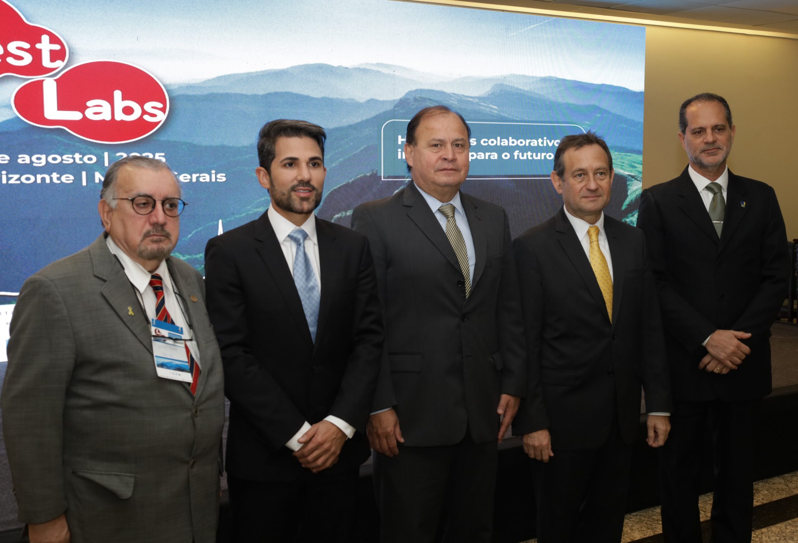 A imagem mostra um grupo de cinco homens, todos de terno e gravata, posando para uma foto em um evento.