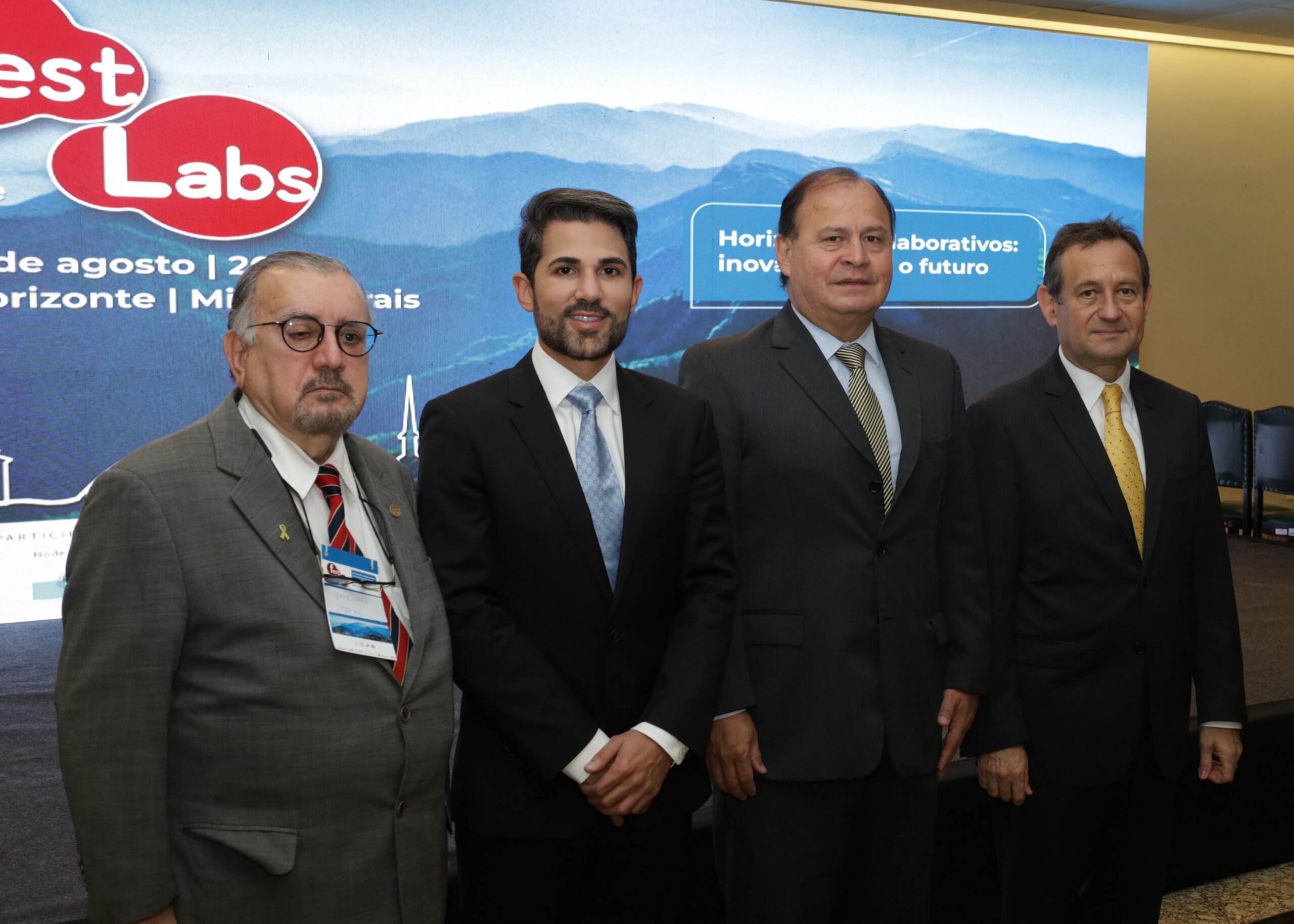 A imagem mostra quatro homens, todos de terno e gravata, posando para uma foto em um evento.