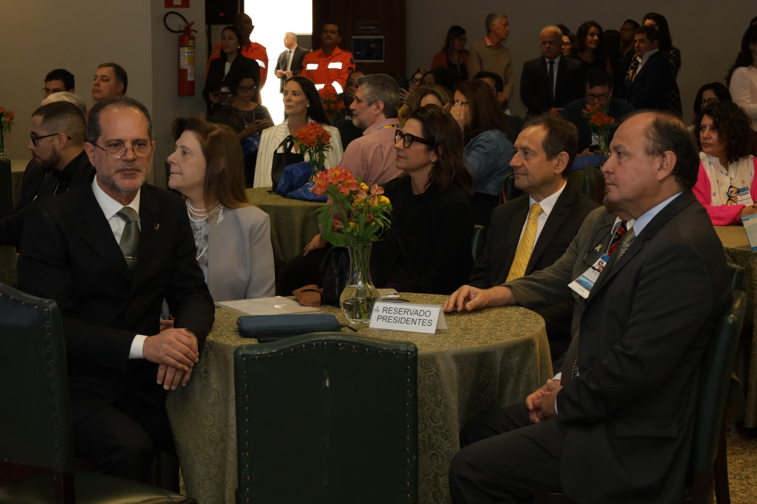 A imagem mostra várias pessoas em trajes formais sentadas em mesas redondas em um evento.