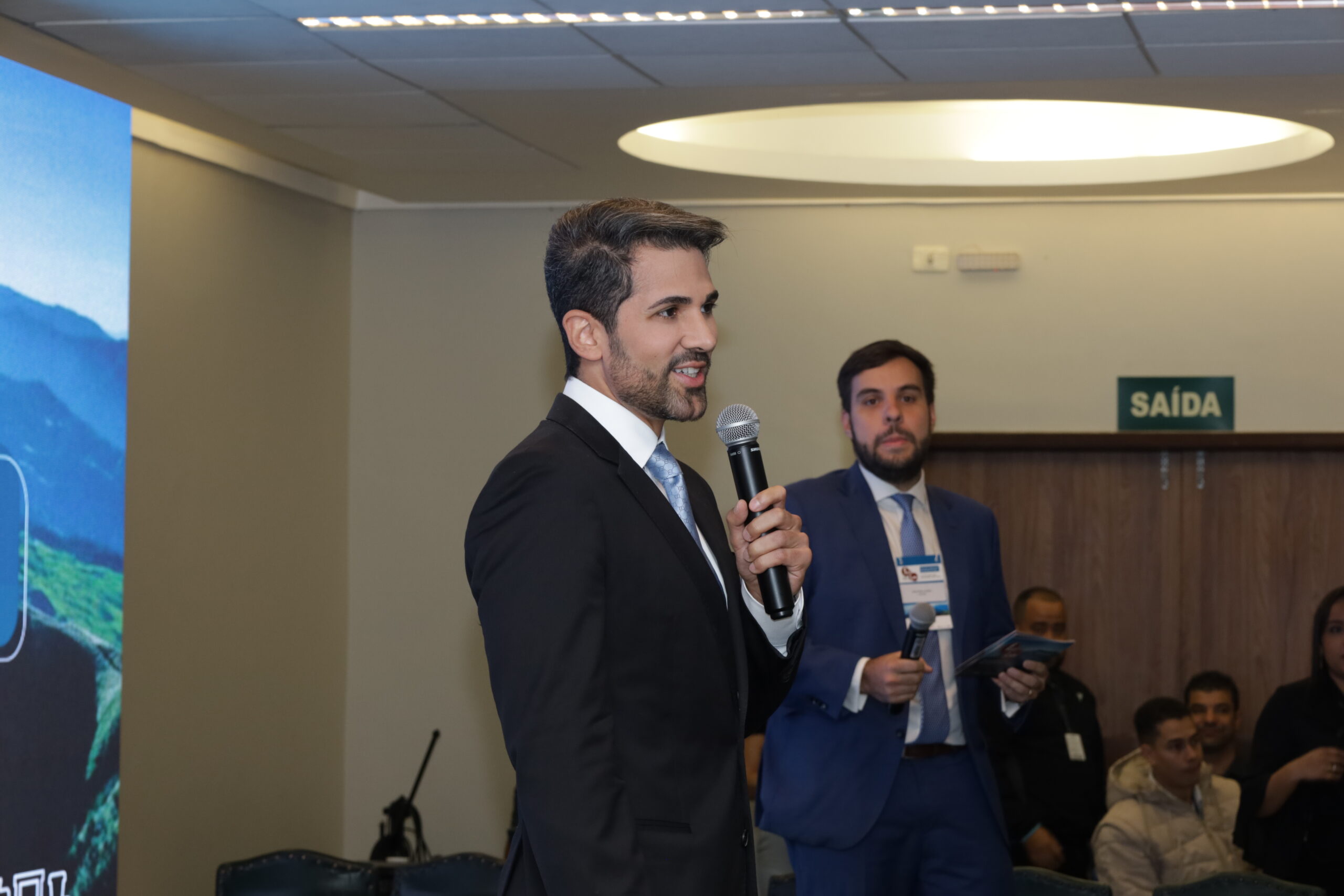 A imagem mostra um homem de terno preto, camisa branca e gravata azul, em pé e segurando um microfone. Ele tem barba e cabelo escuros, e está sorrindo enquanto fala. Atrás dele, um outro homem com terno azul e gravata azul olha para a frente, com um microfone na mão.