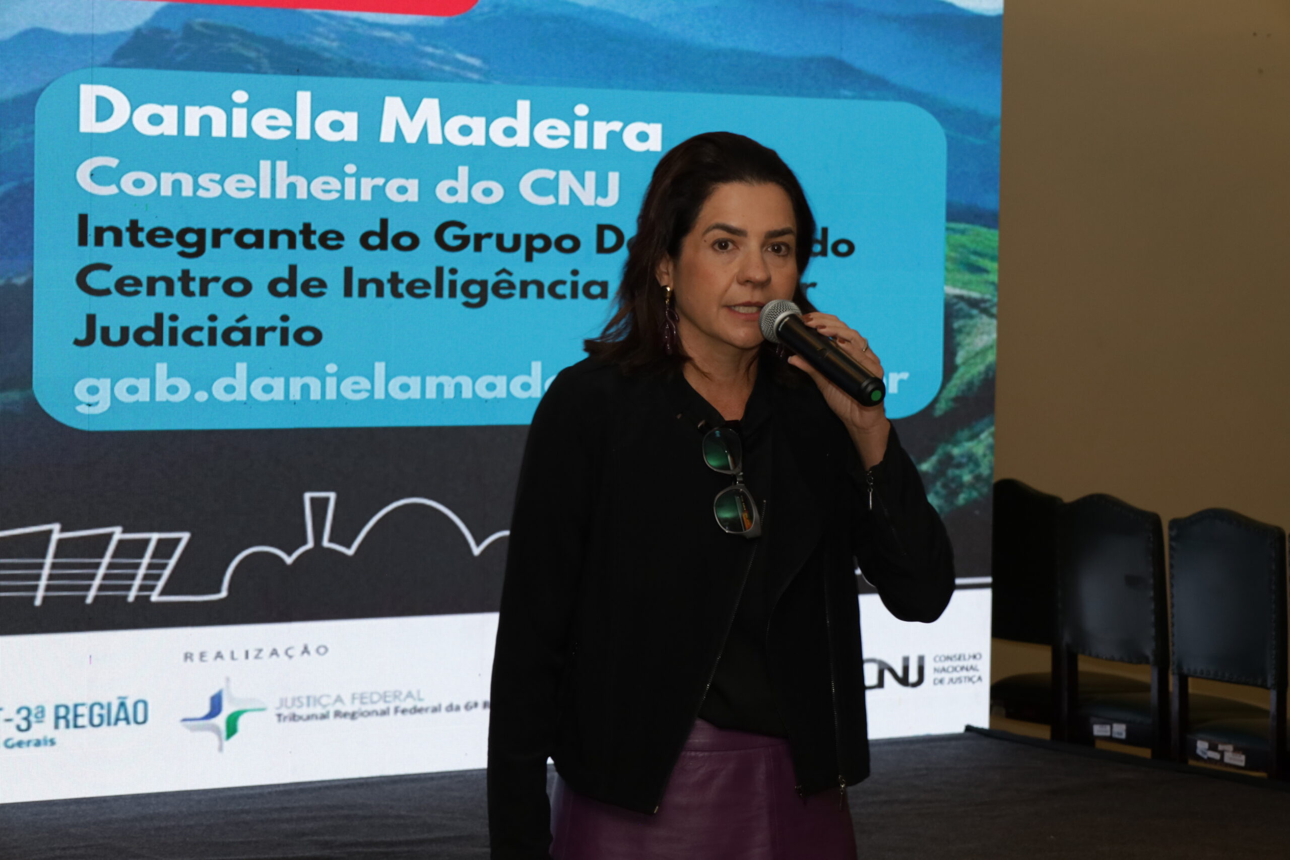 A imagem mostra uma mulher com cabelo escuro, vestindo um blazer preto e uma saia roxa, em pé no palco, falando em um microfone. Ao fundo, um telão exibe um banner azul com seu nome, "Daniela Madeira", e seu cargo, "Conselheira do CNJ".