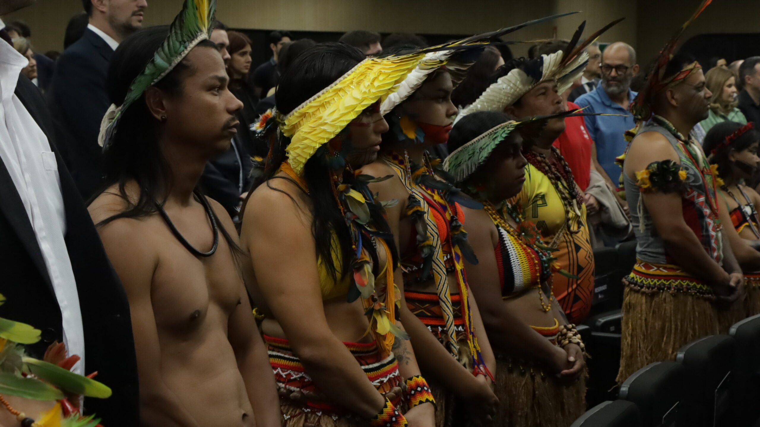 A imagem mostra um grupo de pessoas indígenas em trajes tradicionais, com cocares de penas, colares e adornos coloridos. Eles estão em pé, lado a lado, em uma fileira, em um auditório, com outras pessoas em segundo plano.