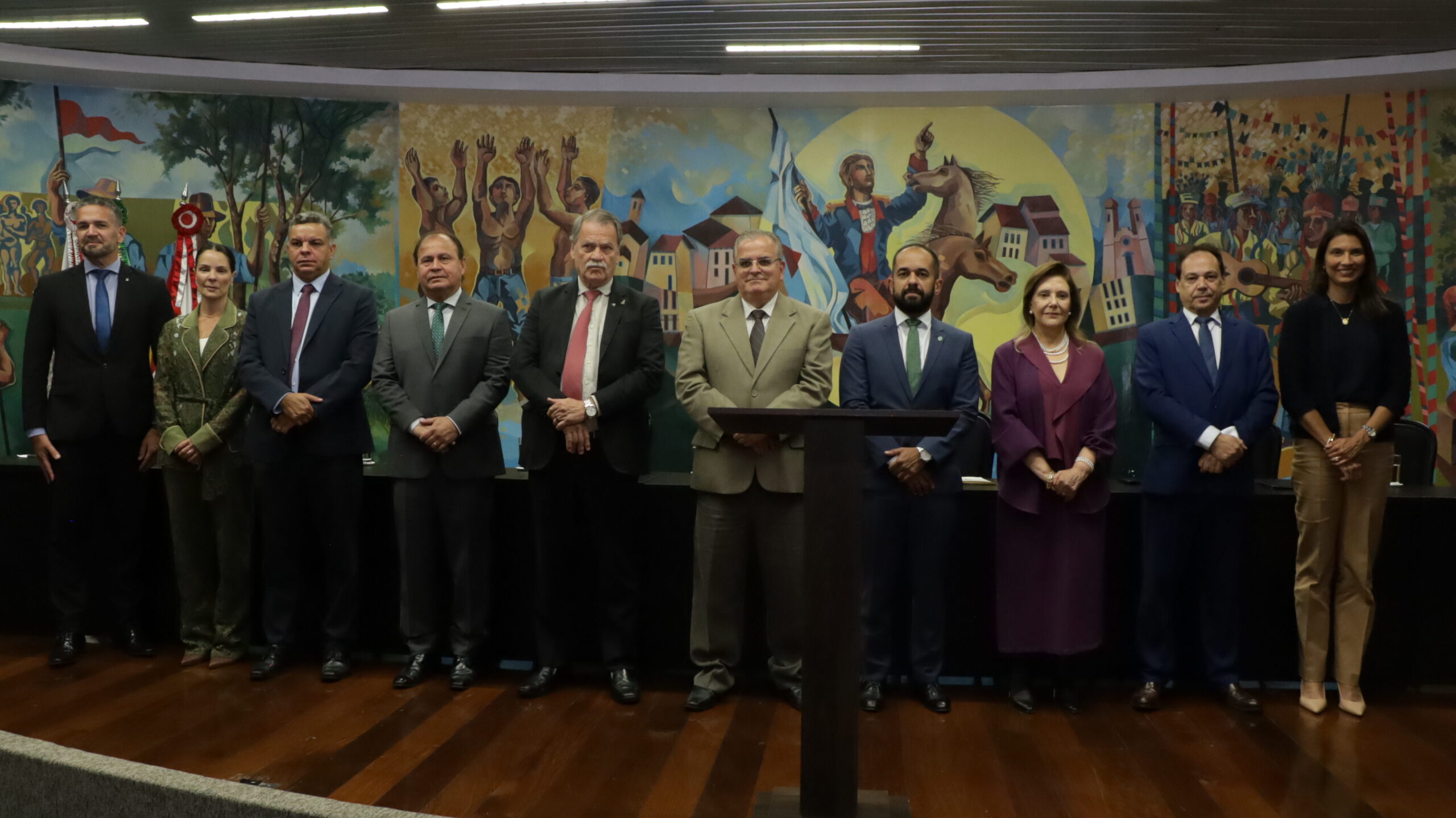 A imagem mostra um grupo de dez pessoas, incluindo homens e mulheres, posando para uma foto em pé, lado a lado, em um ambiente interno. Eles estão em frente a um grande mural pintado, que retrata cenas históricas e culturais. Todos estão em trajes formais. 