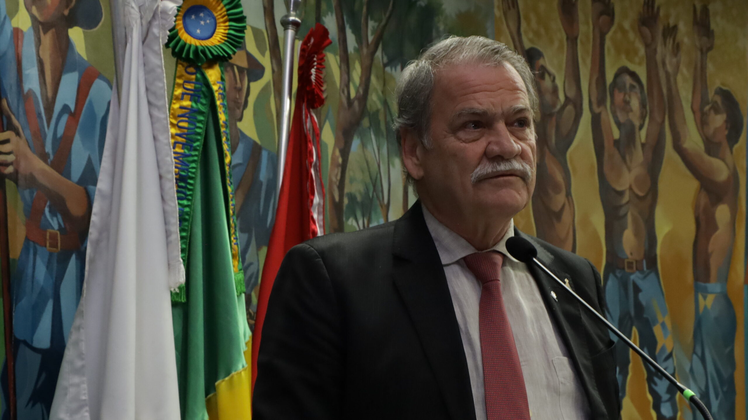 A imagem é um retrato de um homem mais velho, com bigode e bigode grisalhos. Ele está vestindo um terno escuro, uma camisa de cor clara e uma gravata vermelha. O homem está de pé, com um microfone à sua frente.

Ao fundo, há um grande mural pintado com imagens históricas ou folclóricas. Ao lado dele, é possível ver bandeiras hasteadas.