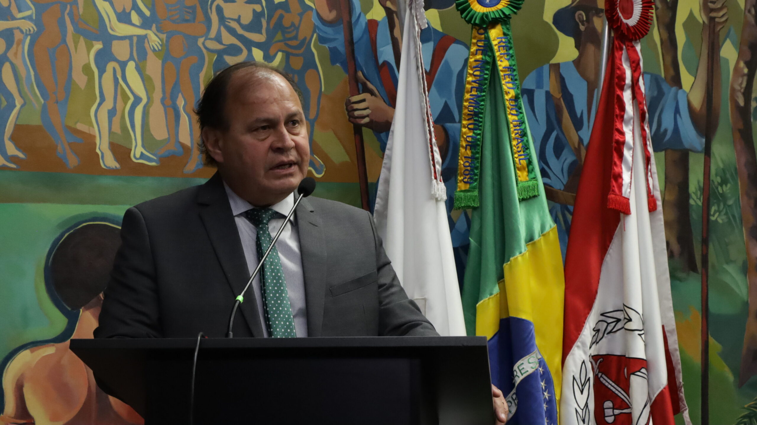 A imagem mostra um homem de meia-idade, com cabelos castanhos e rosto redondo, usando um terno cinza, camisa branca e gravata verde com pequenos pontos brancos. Ele está atrás de um púlpito escuro e parece estar discursando, com a boca aberta em um gesto de fala. À sua direita, há uma fileira de bandeiras hasteadas.