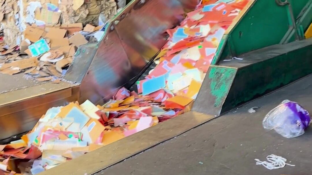 A imagem mostra uma grande quantidade de resíduos plásticos coloridos e papelão em uma esteira transportadora. Há pedaços de plástico de diversas cores, como laranja, azul, rosa e branco, descendo por uma rampa metálica. Ao lado da esteira, é possível ver um pedaço de plástico roxo e branco. Ao fundo, uma grande pilha de papelão.