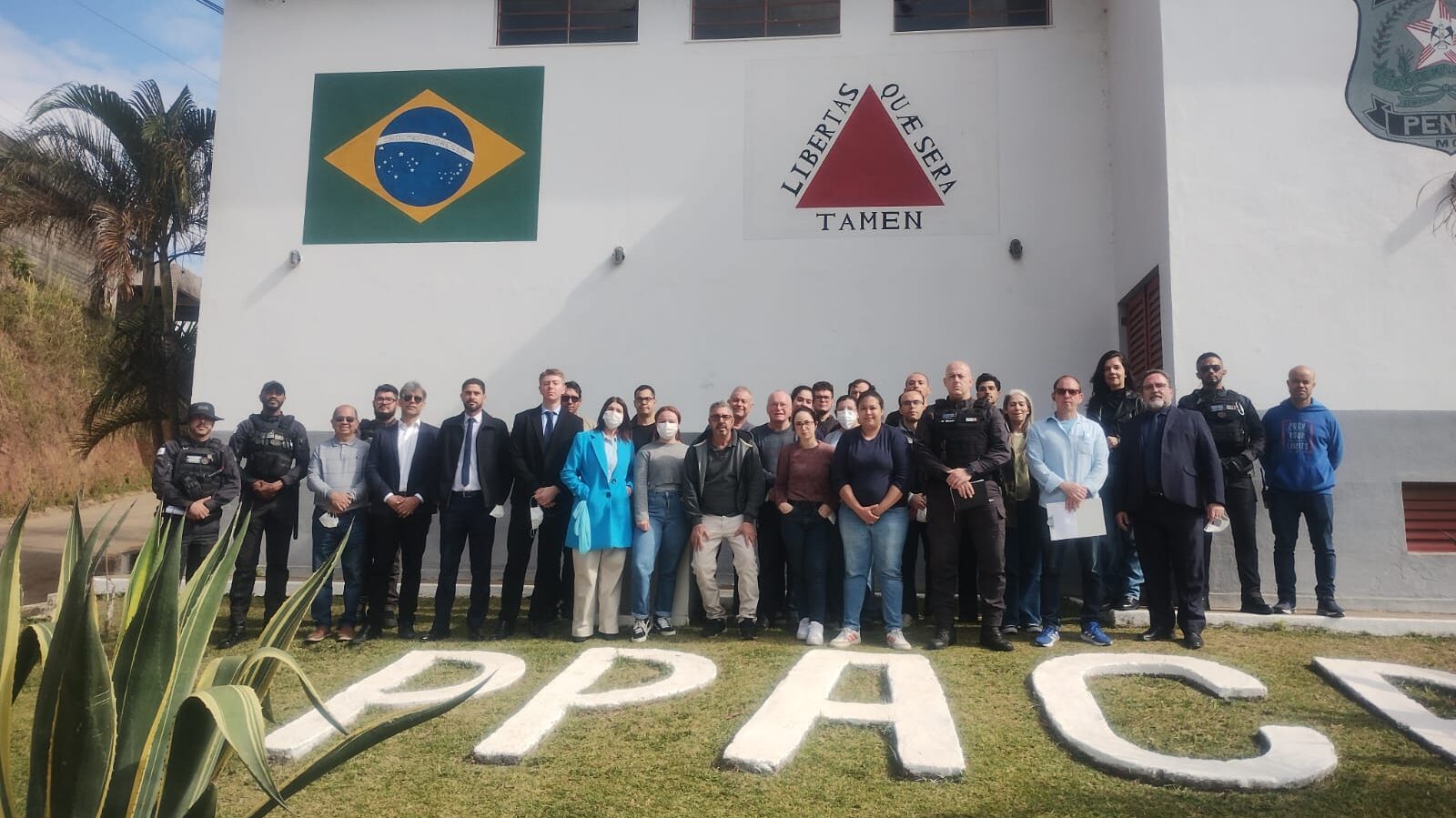 Esta imagem mostra um grupo de pessoas, incluindo policiais e autoridades, em frente a um prédio. Atrás do grupo, na parede do edifício, há uma bandeira do Brasil e de Minas Gerais na parede. A foto foi tirada em um dia claro.