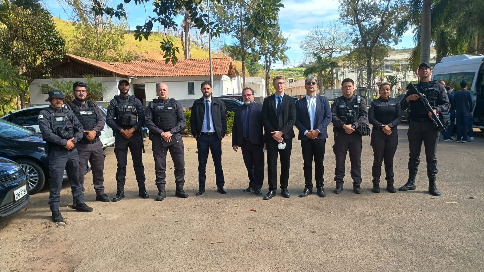A imagem mostra um grupo de pessoas em frente a uma área arborizada. O grupo é composto por policiais, que usam uniformes pretos, coletes e equipamentos táticos. Alguns deles seguram fuzis. Há também quatro homens que parecem ser civis, vestidos com trajes sociais, como ternos e camisas. Eles estão de pé, lado a lado, posando para a foto. No fundo, é possível ver um prédio, algumas árvores e carros estacionados.