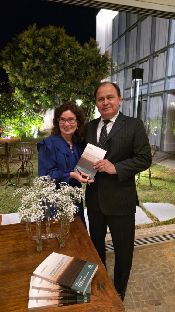 A imagem mostra duas pessoas, um homem e uma mulher, posando para uma foto em um jardim de inverno, com iluminação noturna. A mulher, de cabelo castanho encaracolado e óculos, usa uma blusa azul-escura com babados. Ela está segurando um livro.

Ao lado dela, o homem está de terno escuro, camisa branca e gravata. Ele também segura o livro. No primeiro plano, uma mesa de madeira tem uma pilha de cópias do mesmo livro, com flores brancas em vasos ao lado. O ambiente é iluminado e parece ser um evento de lançamento de livro ou uma celebração. O livro tem o título "Direito & Economia Comportamental: Aspectos jurídicos e filosóficos do poder judicial".