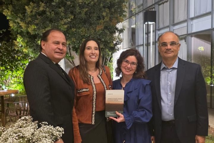 A imagem mostra um grupo de quatro pessoas posando para uma foto em um ambiente interno que se parece com uma sala de eventos ou um jardim de inverno, com plantas e iluminação noturna. Duas mulheres e dois homens estão em pé, lado a lado. A mulher no centro do grupo segura um livro.