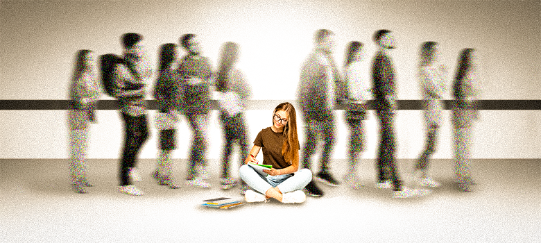 A imagem mostra uma jovem sentada no chão, de pernas cruzadas, com um caderno e uma caneta na mão. Ela usa óculos e uma camiseta marrom e sorri levemente. Ao redor dela, um grupo de pessoas está em pé e borrado, como se estivessem em movimento.