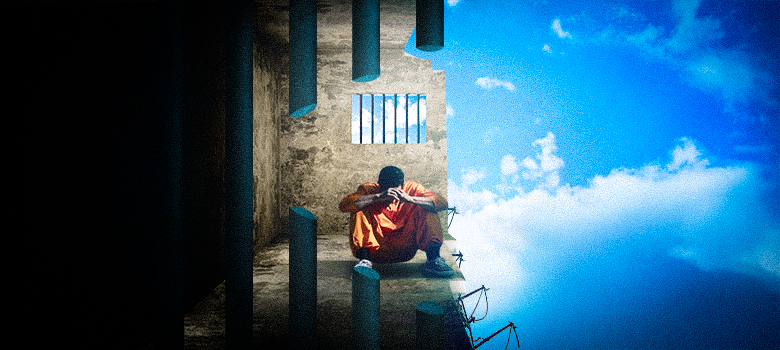 A imagem é uma montagem que retrata uma cena de confinamento e liberdade. No lado esquerdo, um homem de macacão laranja está sentado no chão de uma cela, com a cabeça baixa e as mãos apoiadas nos joelhos. Ele está em uma cela cinza e escura, com uma pequena janela gradeada. A cela tem paredes e um teto de concreto, com uma luz vindo da janela.

O lado direito da imagem é dominado por um céu azul com nuvens brancas e claras, representando a liberdade e o mundo exterior. Uma linha vertical divide as duas cenas, simbolizando a separação entre a prisão e a liberdade. No lado direito, há a ponta de uma balança da justiça, que faz alusão ao sistema judicial.