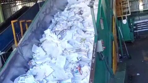 A imagem mostra uma grande quantidade de papel branco em uma esteira transportadora em uma instalação industrial. O papel está amontoado e sendo transportado por uma rampa metálica verde. Ao fundo, estruturas metálicas e outros equipamentos industriais são visíveis.
