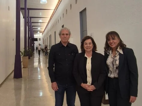 A imagem mostra três pessoas posando para uma foto em um corredor longo e iluminado. No centro, uma mulher de meia-idade com cabelos castanhos escuros e ondulados sorri para a câmera.
À sua esquerda, um homem com cabelos grisalhos e cacheados, de meia-idade, veste uma camisa preta de mangas compridas e calça jeans escura. À direita da mulher ao centro, uma mulher de cabelos castanhos e curtos, e usa um blazer azul-marinho sobre uma blusa estampada em preto e branco.
O corredor tem paredes brancas e pilares roxos no lado esquerdo, com o chão em azulejos cinzas e brilhantes. Outras pessoas podem ser vistas ao fundo do corredor.