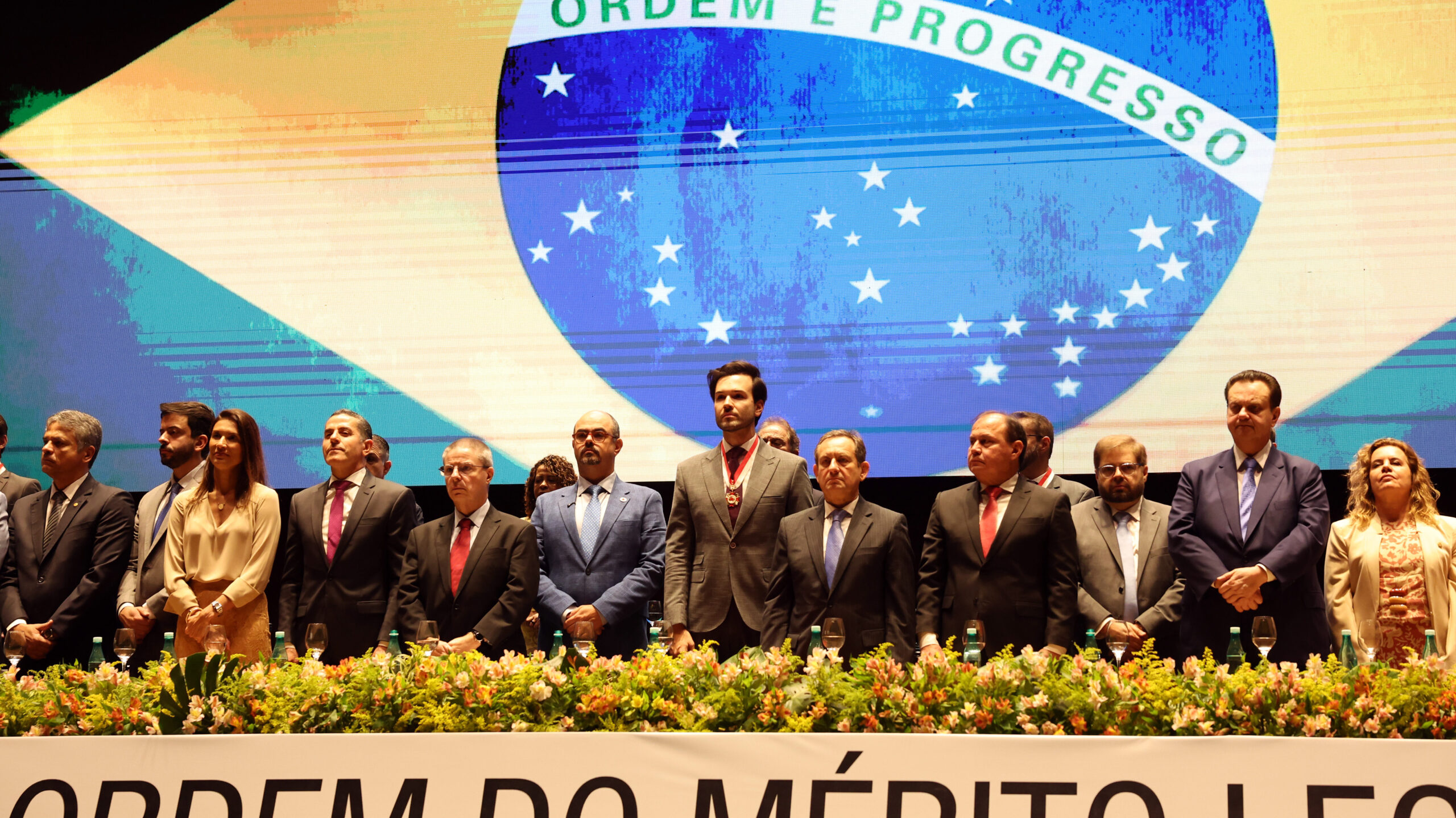 A imagem retrata um grupo de pessoas em pé em uma fileira durante uma cerimônia formal. Todos estão vestidos com trajes sociais. Eles estão atrás de uma grande jardineira com folhagens verdes e algumas flores, que se estende por toda a frente.
No fundo, há um painel ou tela gigante exibindo a bandeira do Brasil em alta resolução, com o dístico "ORDEM E PROGRESSO" claramente visível.