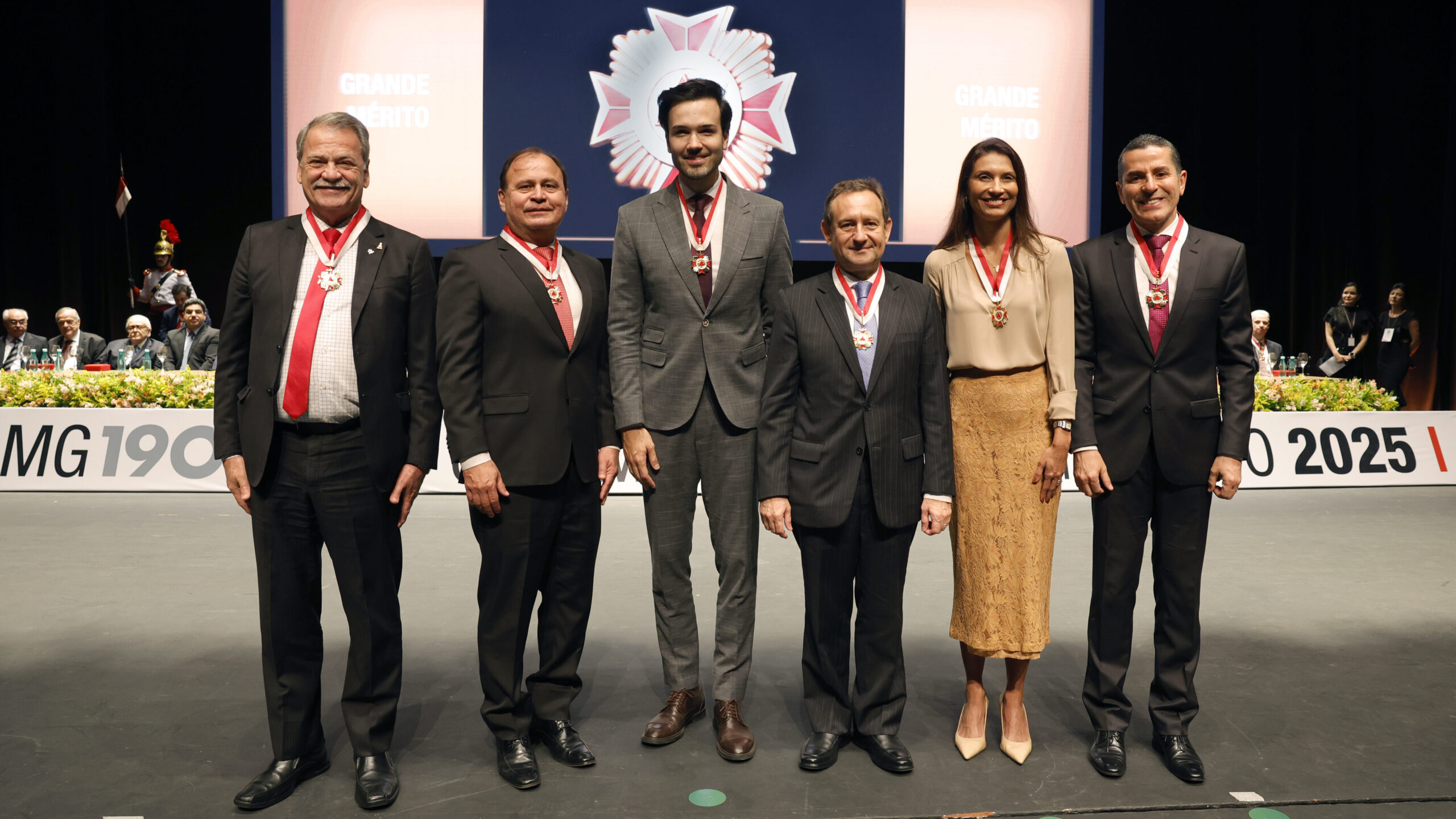 A fotografia registra um momento de uma cerimônia formal, onde seis pessoas (cinco homens e uma mulher) estão em pé e alinhadas em um palco, todas condecoradas com medalhas de prestígio.