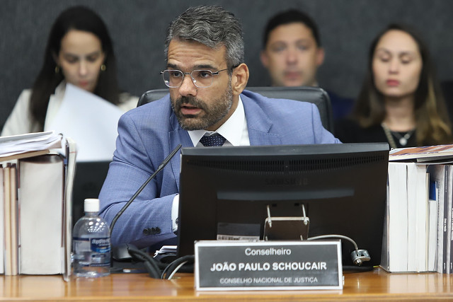 A imagem foca em um homem sentado à frente de uma mesa de madeira, usando um terno azul claro, óculos e barba. Ele está olhando para cima e à sua esquerda, com a boca ligeiramente aberta. Em sua frente, há um monitor de computador com uma placa de identificação que lê "Conselheiro JOÃO PAULO SHOUCAIR CONSELHO NACIONAL DE JUSTIÇA". Ao lado do monitor, há pilhas de documentos e uma garrafa de água. Ao fundo, outras pessoas desfocadas estão sentadas em uma mesa.