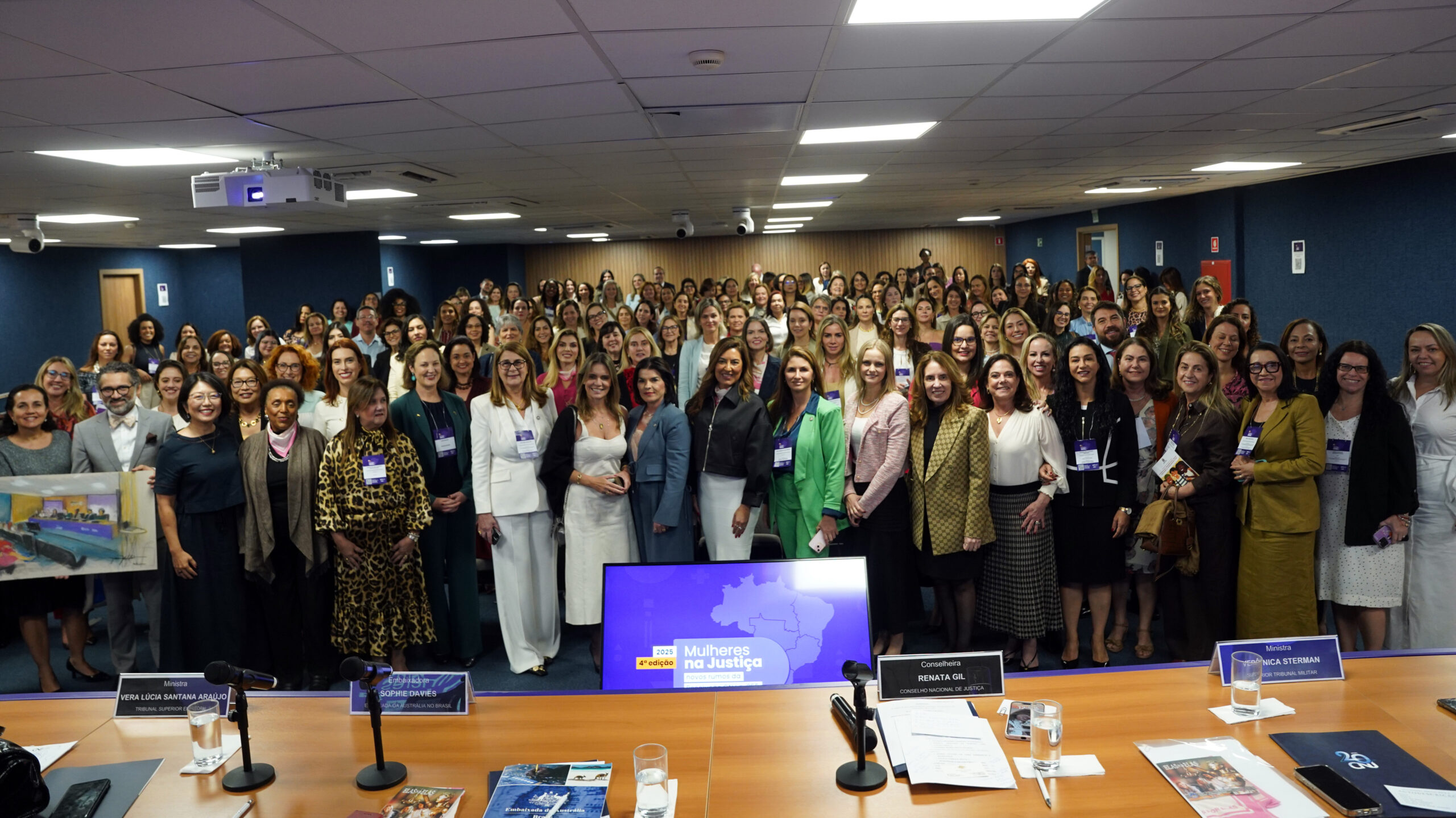 Foto panorâmica de um evento, mostrando um grande grupo de mulheres (e alguns poucos homens) em pé, posando para uma foto de grupo em uma sala de conferências.

A grande maioria das pessoas está vestida com trajes formais e casuais-chiques.

No centro do primeiro plano, há um monitor de LED exibindo o tema do evento: "Mulheres na Justiça" com um mapa do Brasil em roxo.

O ambiente possui paredes azuis escuras e o primeiro plano é ocupado por uma mesa de conferência de madeira clara com microfones, água e cartões de identificação. Tags de identificação são visíveis em algumas pessoas da frente.