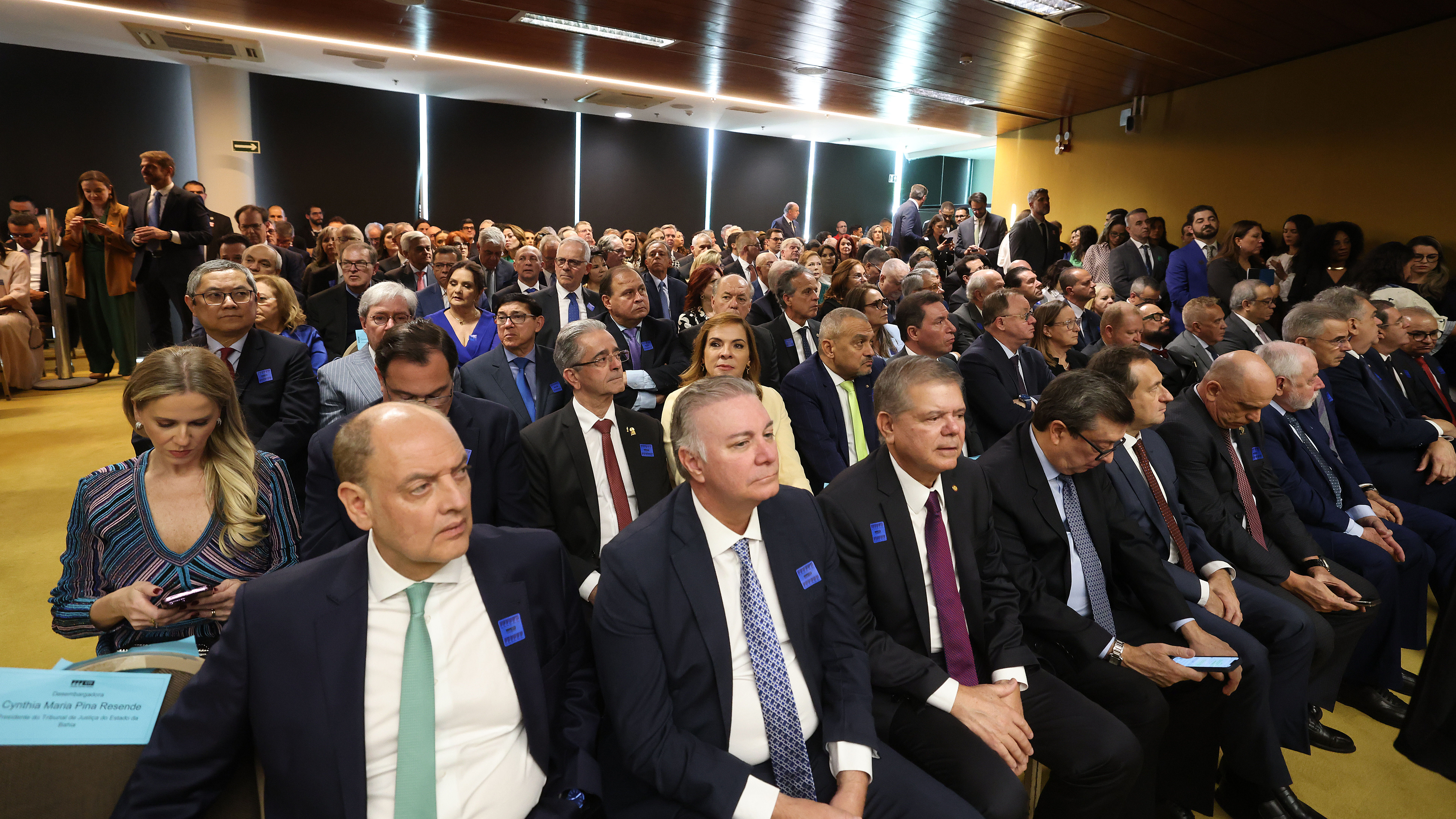 Uma foto de uma grande audiência ou evento lotado, com dezenas de pessoas sentadas em fileiras de cadeiras. A maioria veste trajes sociais escuros (ternos), e algumas pessoas estão olhando para o lado. O ambiente tem paredes claras e um teto de madeira.
