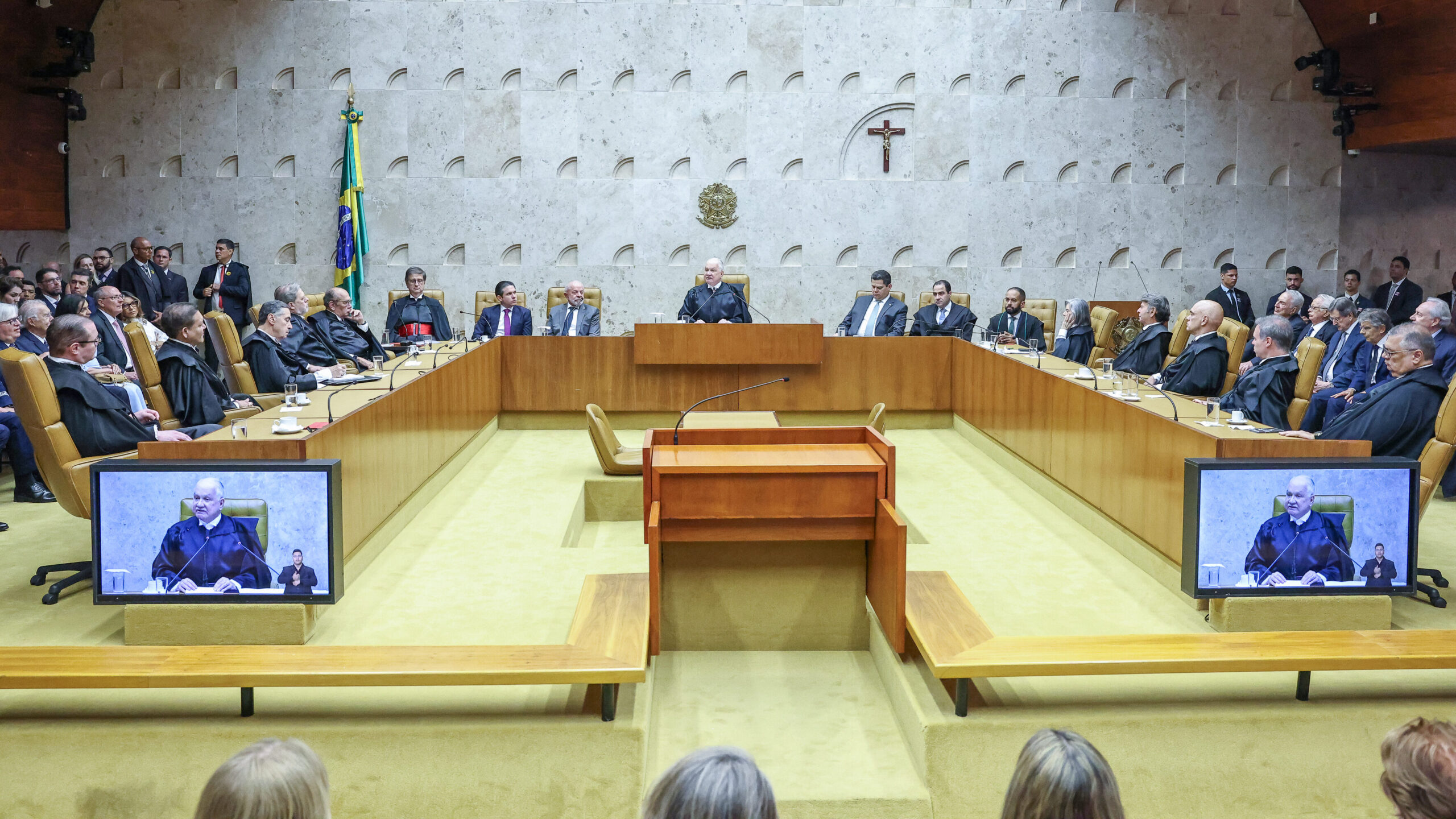 Vista de uma sala de audiência ou plenário, com vários homens de toga e terno sentados em uma longa mesa em formato de "U", voltados para o centro onde há uma tribuna de madeira. A cena é gravada e transmitida em duas telas de televisão dispostas no chão, e há a presença de bandeiras e um crucifixo na parede de fundo.