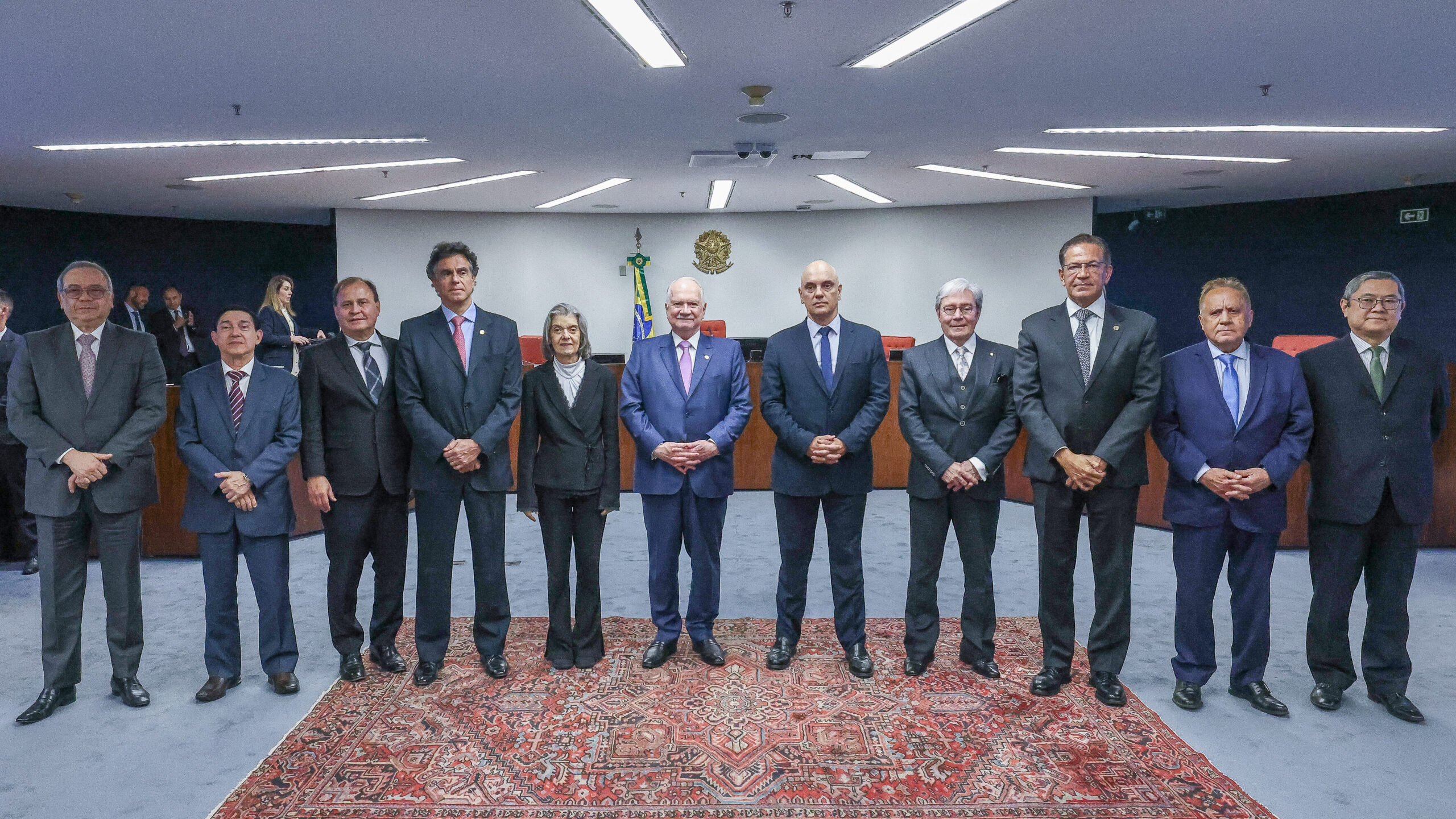 Um grupo de onze pessoas (dez homens e uma mulher) está posando em pé em uma sala formal de plenário ou conferência. Todos vestem trajes formais, a maioria ternos escuros.

O grupo está alinhado em frente a um tapete vermelho e estampado. Ao centro, há uma planta com a bandeira do Brasil. O fundo é uma parede escura e lisa, com o teto de madeira ripada e iluminação embutida. Há outros participantes sentados ao fundo e nas laterais da sala.