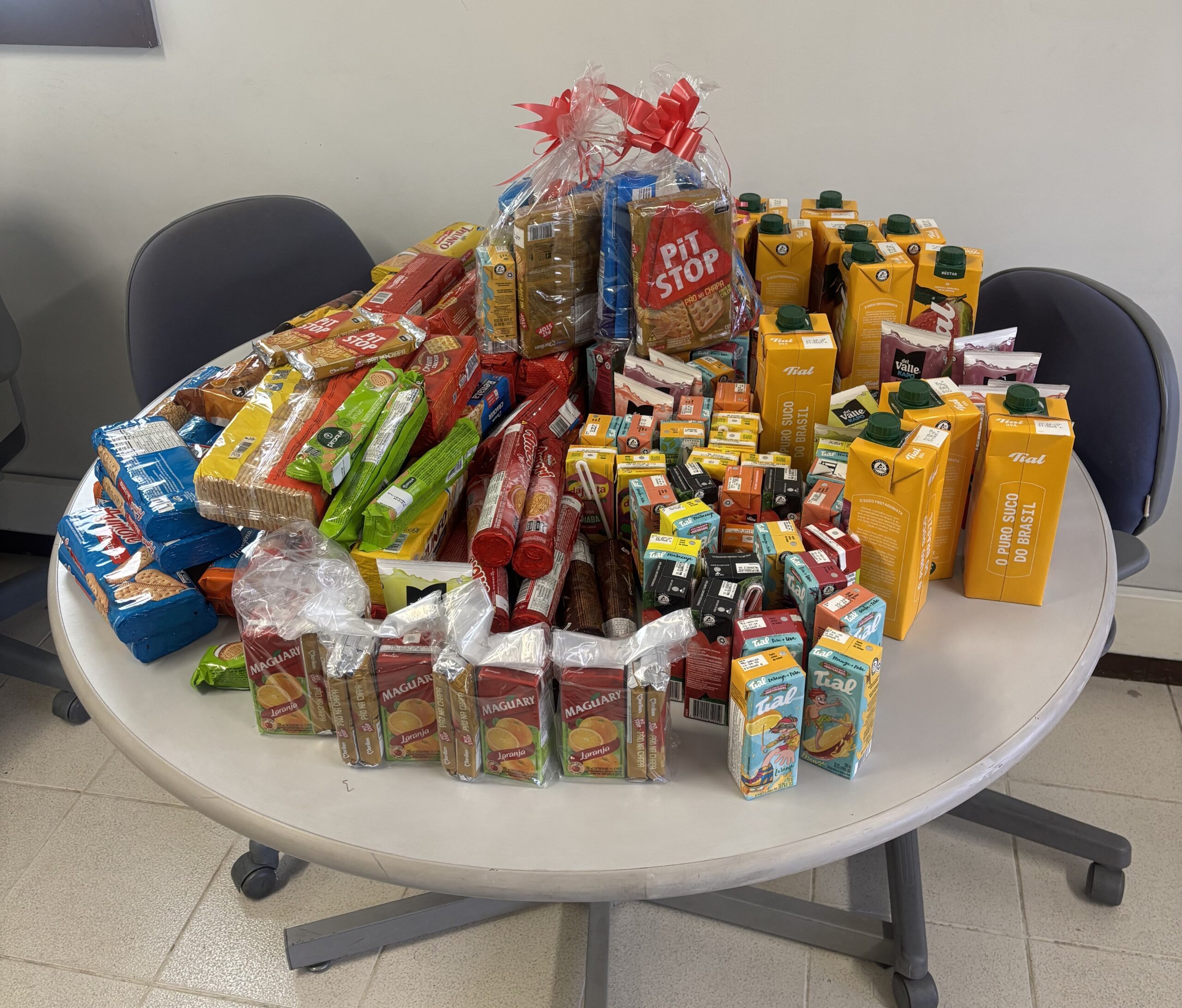 A imagem mostra uma grande quantidade de alimentos não perecíveis dispostos sobre uma mesa redonda. Há muitos biscoitos e salgadinhos em embalagens de diversas cores, além de diversas caixinhas de suco. No centro, há uma embalagem maior de biscoitos, embrulhada em um laço vermelho. Os itens estão organizados de forma a parecerem doações.