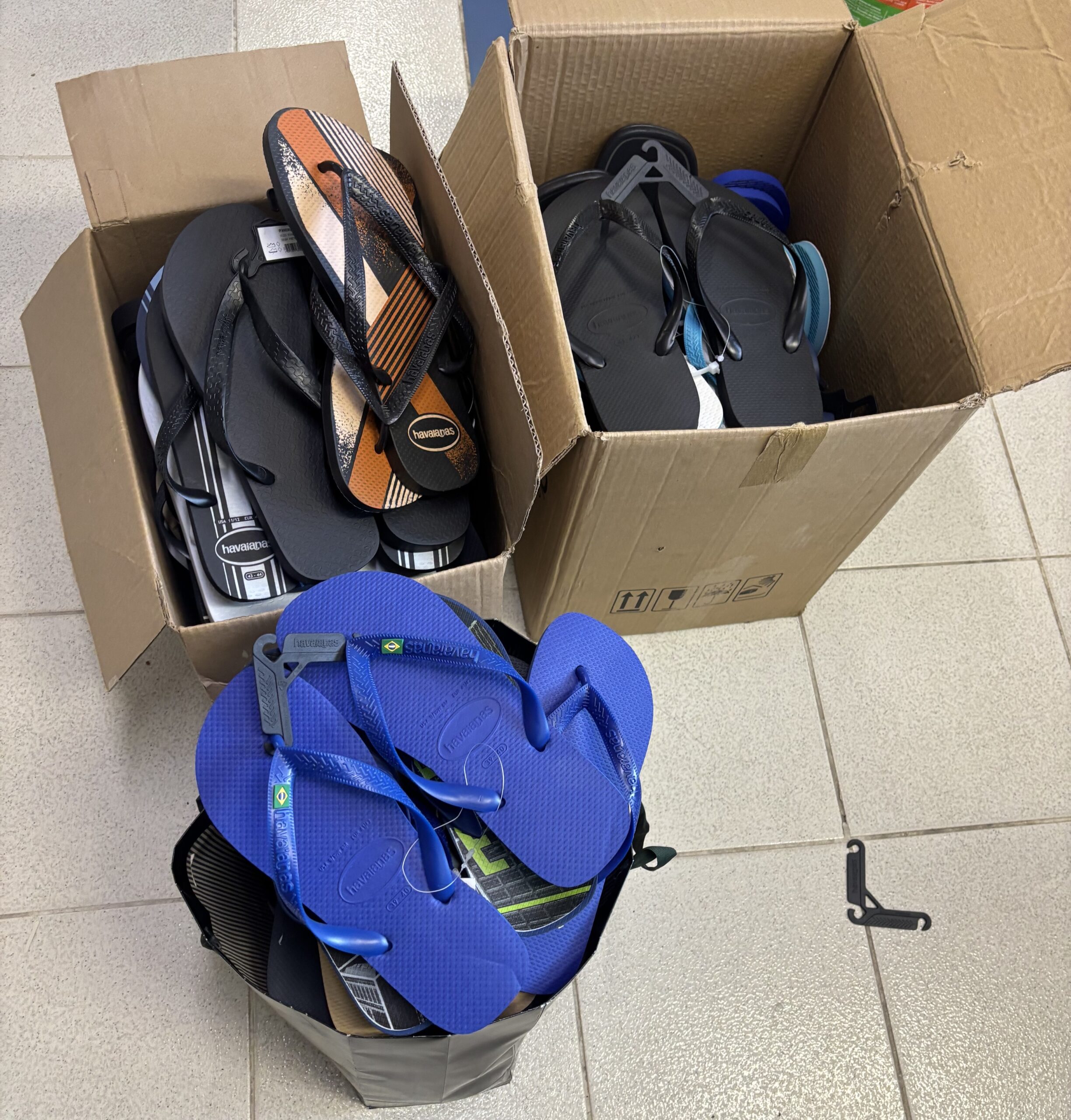 A imagem mostra três recipientes cheios de sandálias tipo chinelo. À frente, um saco preto está cheio de chinelos azuis. Também há duas caixas de papelão abertas, repletas de chinelos pretos e de outras cores e modelos.