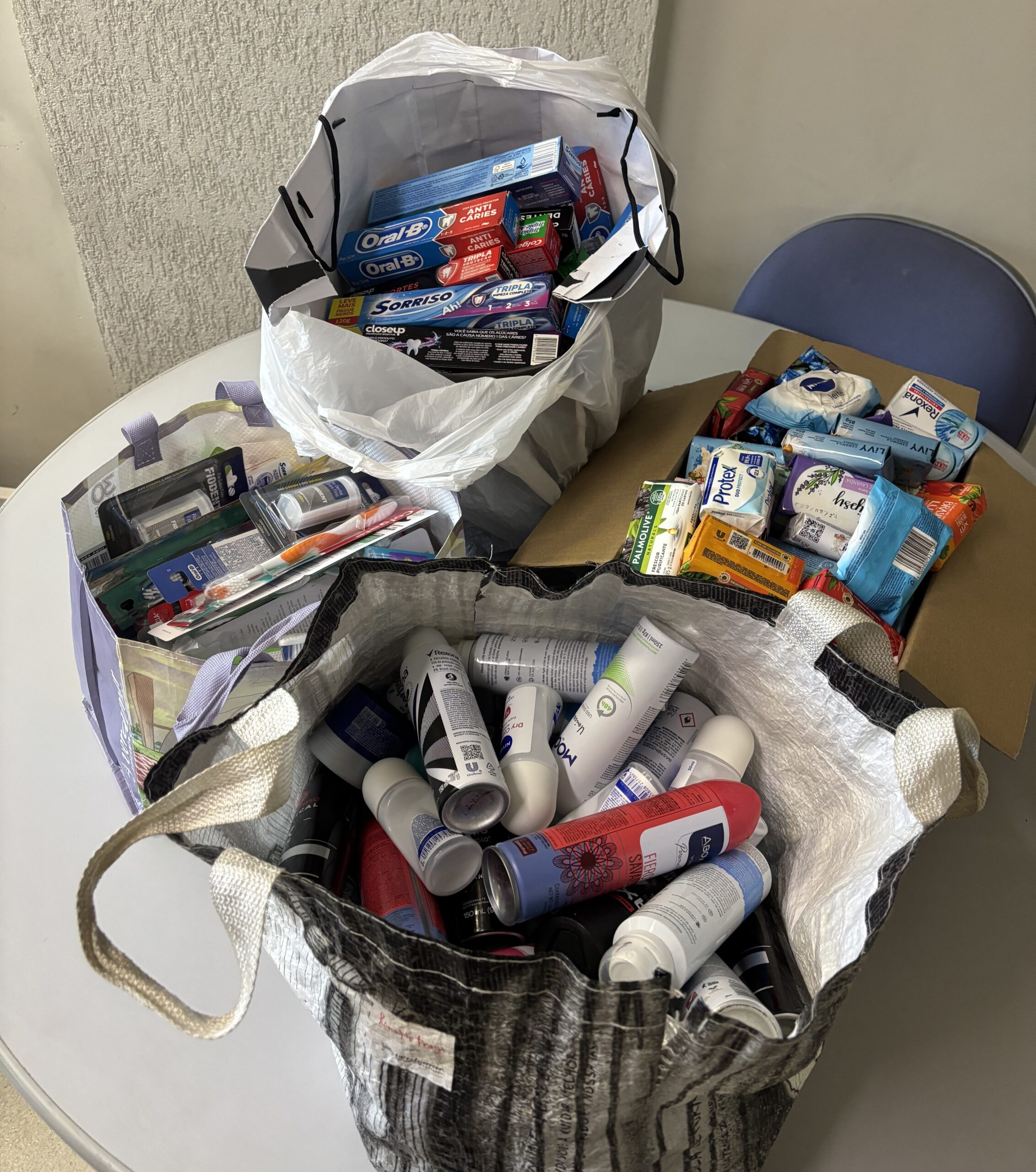 A foto, tirada de cima, mostra três sacolas e uma caixa cheias de produtos de higiene pessoal, como desodorantes em spray e roll-on, pastas de dente, escovas de dente, sabonetes e embalagens de lenços umedecidos. Os itens estão prontos para doação ou distribuição.