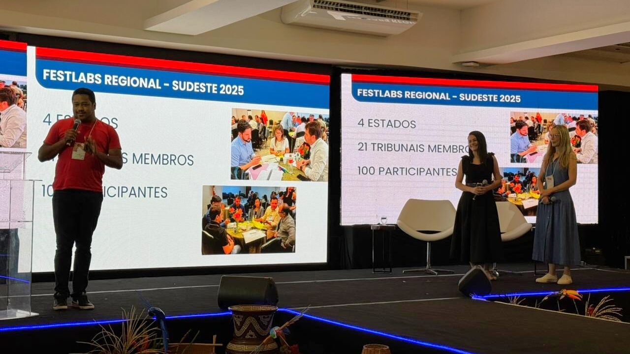 A foto mostra um palco de evento com duas telas grandes projetando o mesmo conteúdo lado a lado. À esquerda, um homem de camiseta e calça escura está falando ao microfone, de frente para a plateia. À direita, duas mulheres, ambas de vestido, estão em frente à outra tela, segurando microfones.