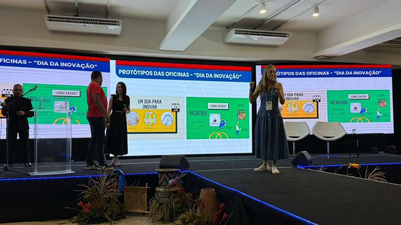 A foto mostra um palco de evento com duas telas grandes exibindo a mesma apresentação. A apresentação contém slides com gráficos, diagramas e textos. No palco, da esquerda para a direita, um homem de terno e gravata está atrás de um púlpito; outro homem de camiseta e calça escura está ao lado de uma mulher de vestido escuro, ambos de costas; e, à direita, outra mulher de vestido comprido, segurando um crachá, está falando e gesticulando. Cadeiras brancas de design moderno estão visíveis no palco.